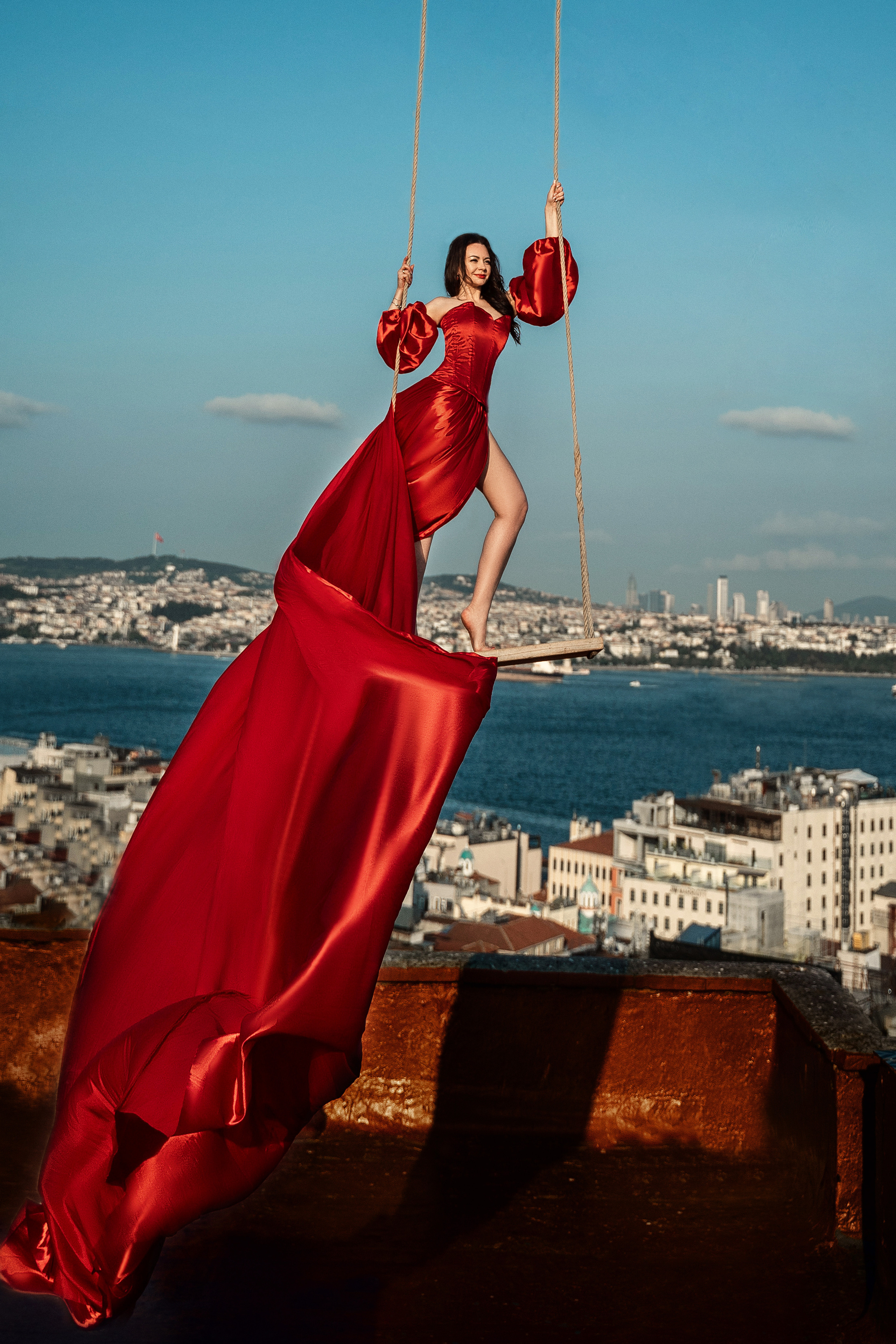 Фотосессия в красном платье с длинным шлейфом на качеле крыши с чайками