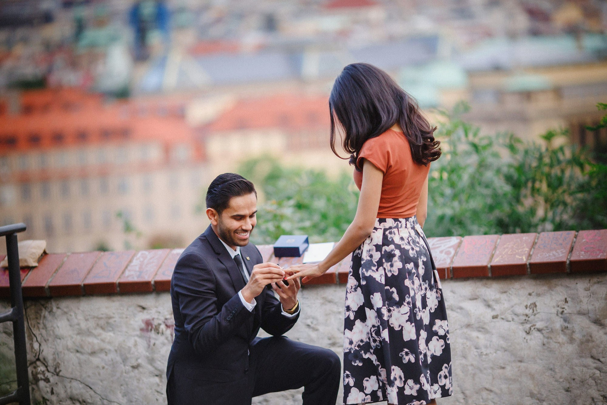 Man smiling while placing engagement ring on partner’s finger high above Prague.