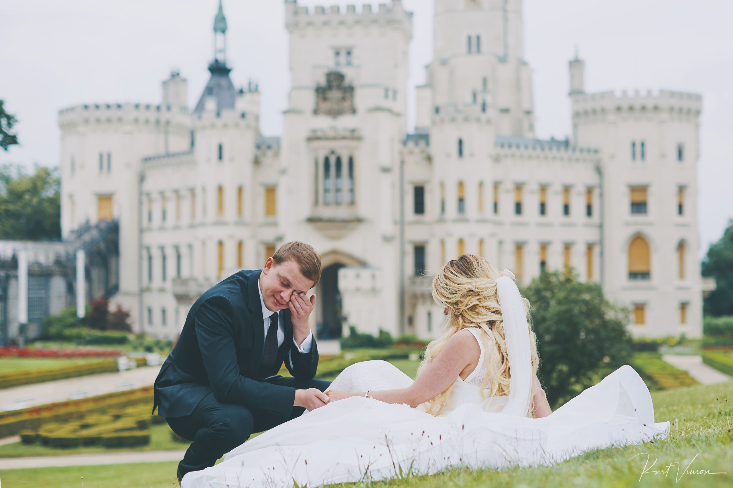A stylishly dressed groom wipes the tears from his eyes after apologizing to his bride for taking a business phone call as she sits patiently on the grounds of the castle where they earlier married.