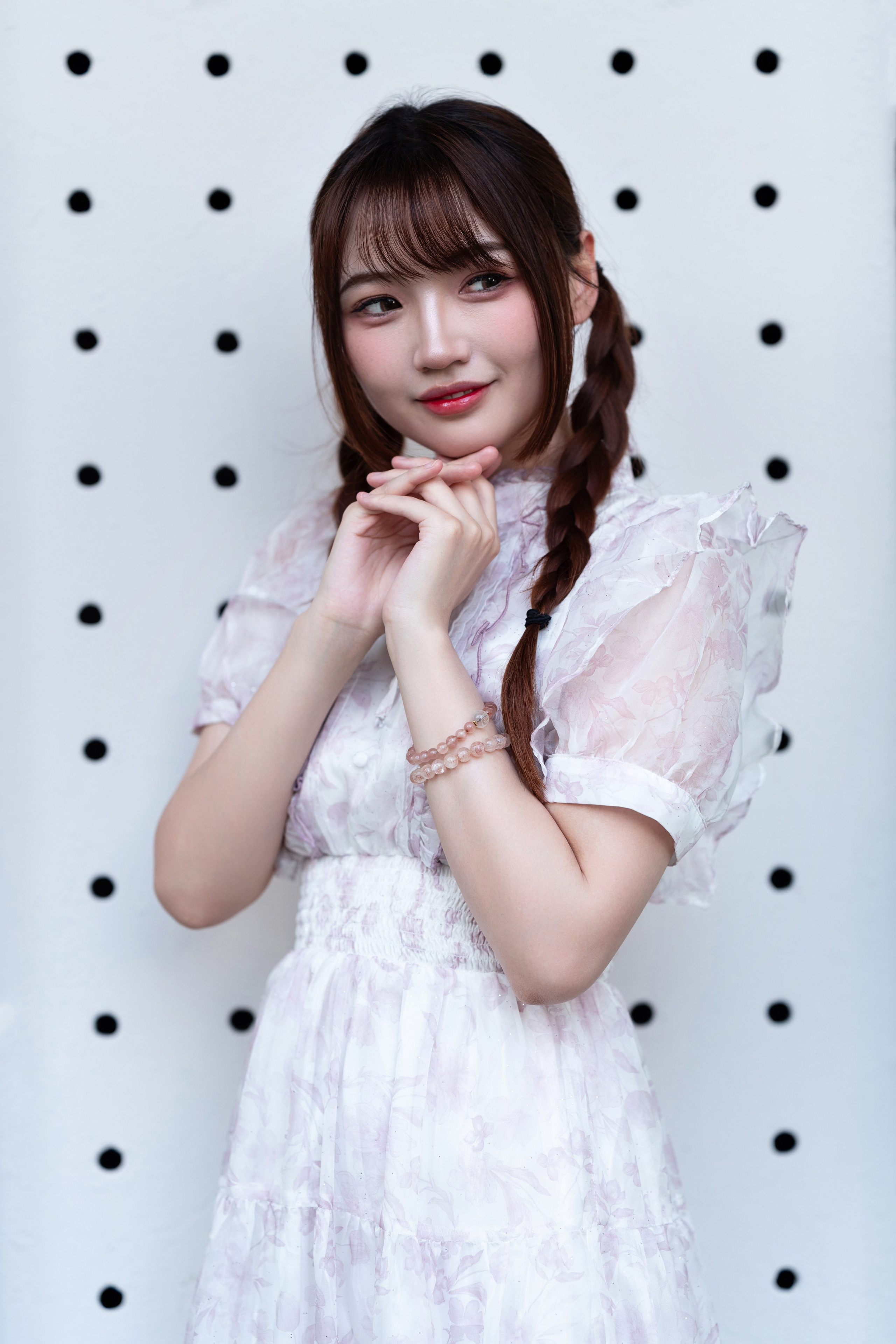 Same model in light-floral dress, hands delicately under chin, posed before a white wall dotted with graphic black holes.
