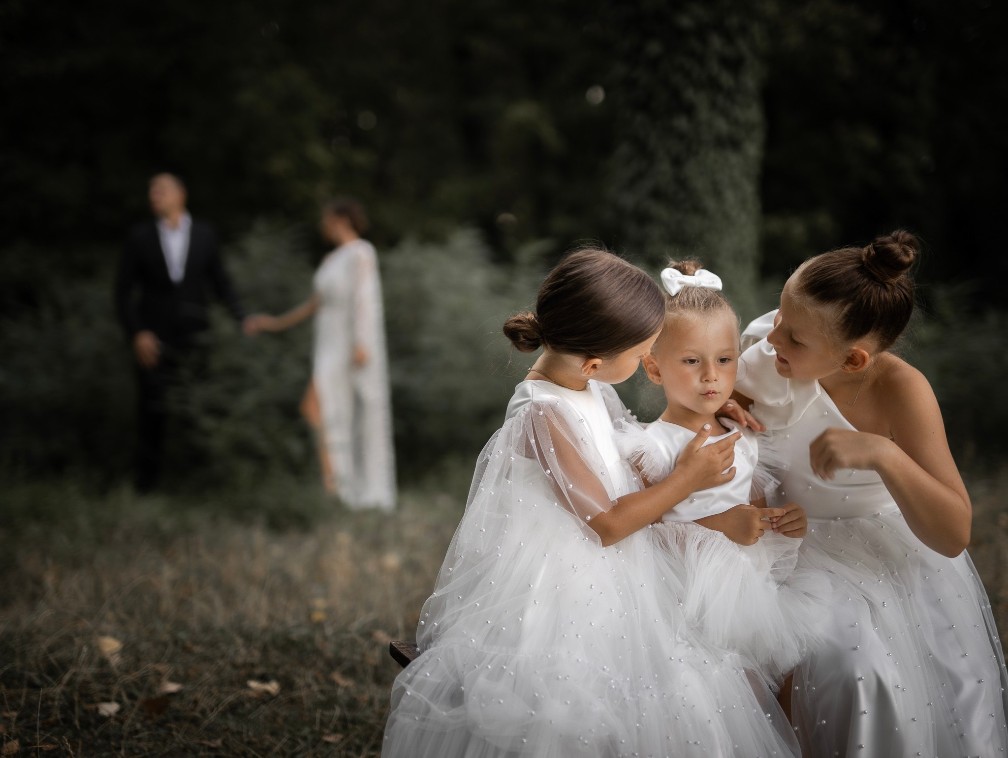 tri male devojčice u belim haljinama pricaju, dok su roditelji u pozadini.