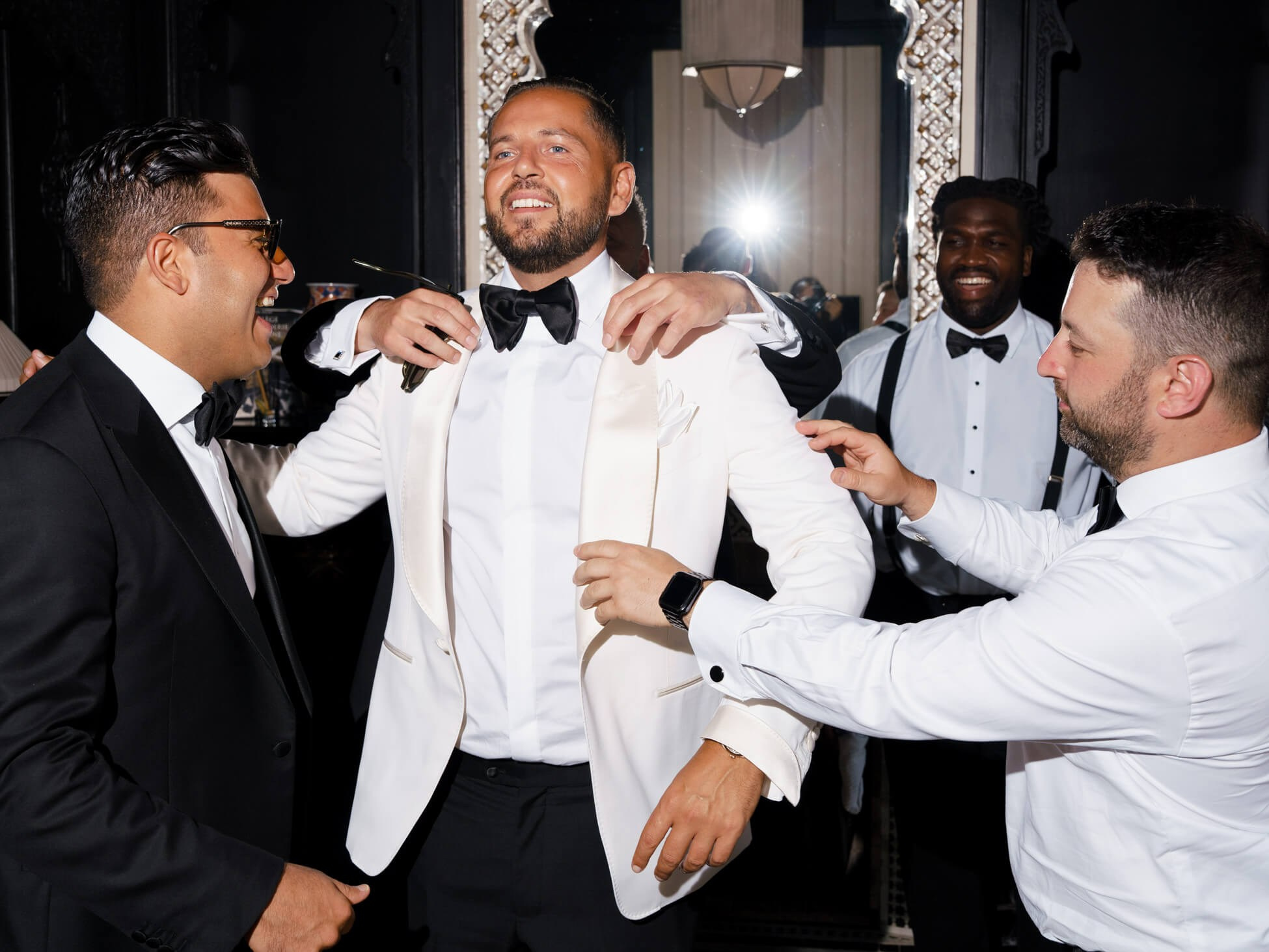 Groomsmen helping the groom into his white tuxedo jacket in a flash-lit candid, destination wedding getting-ready moment in Morocco