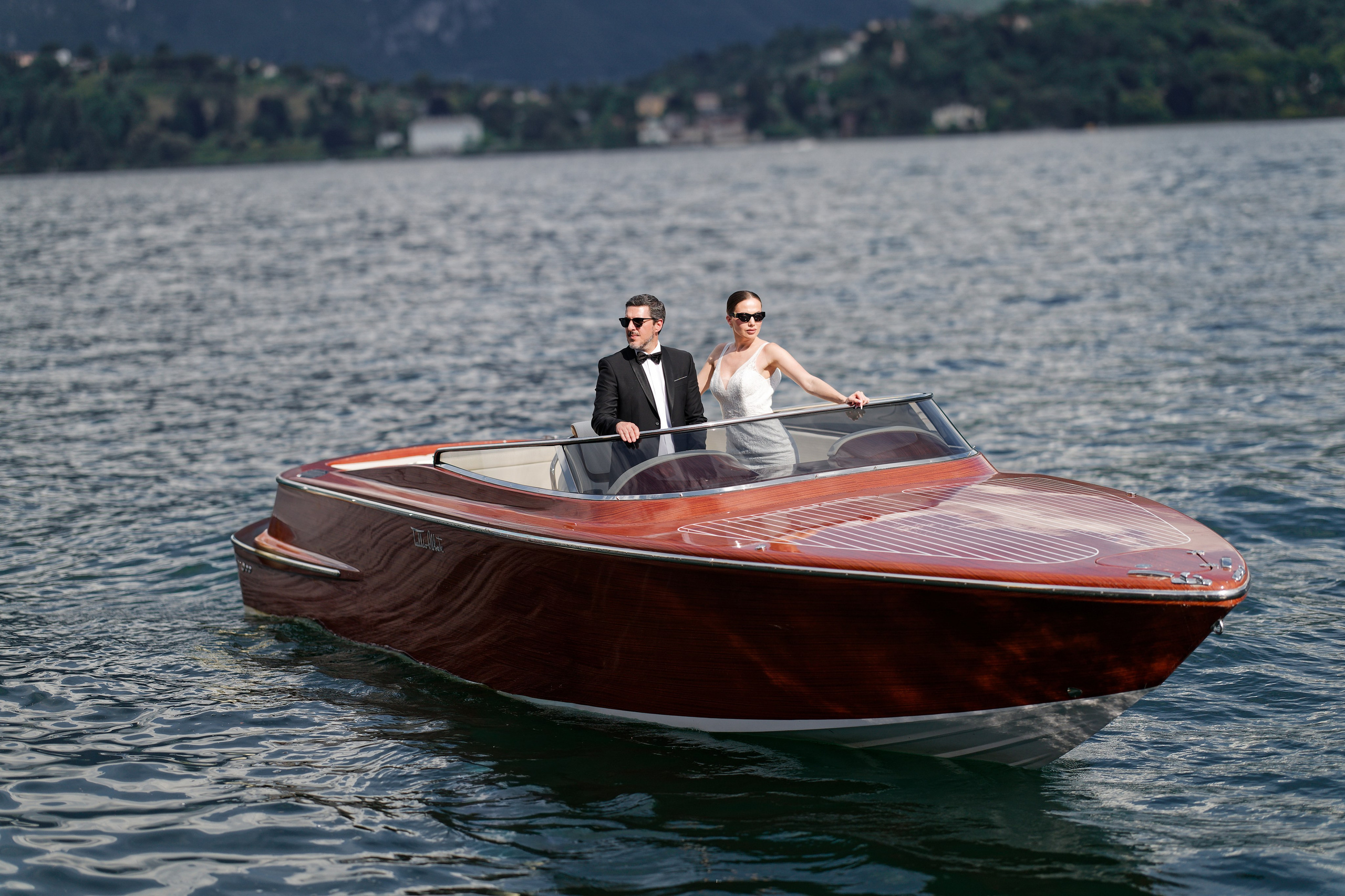 "Wooden boat carrying newlyweds gliding on the water against a mountain backdrop."