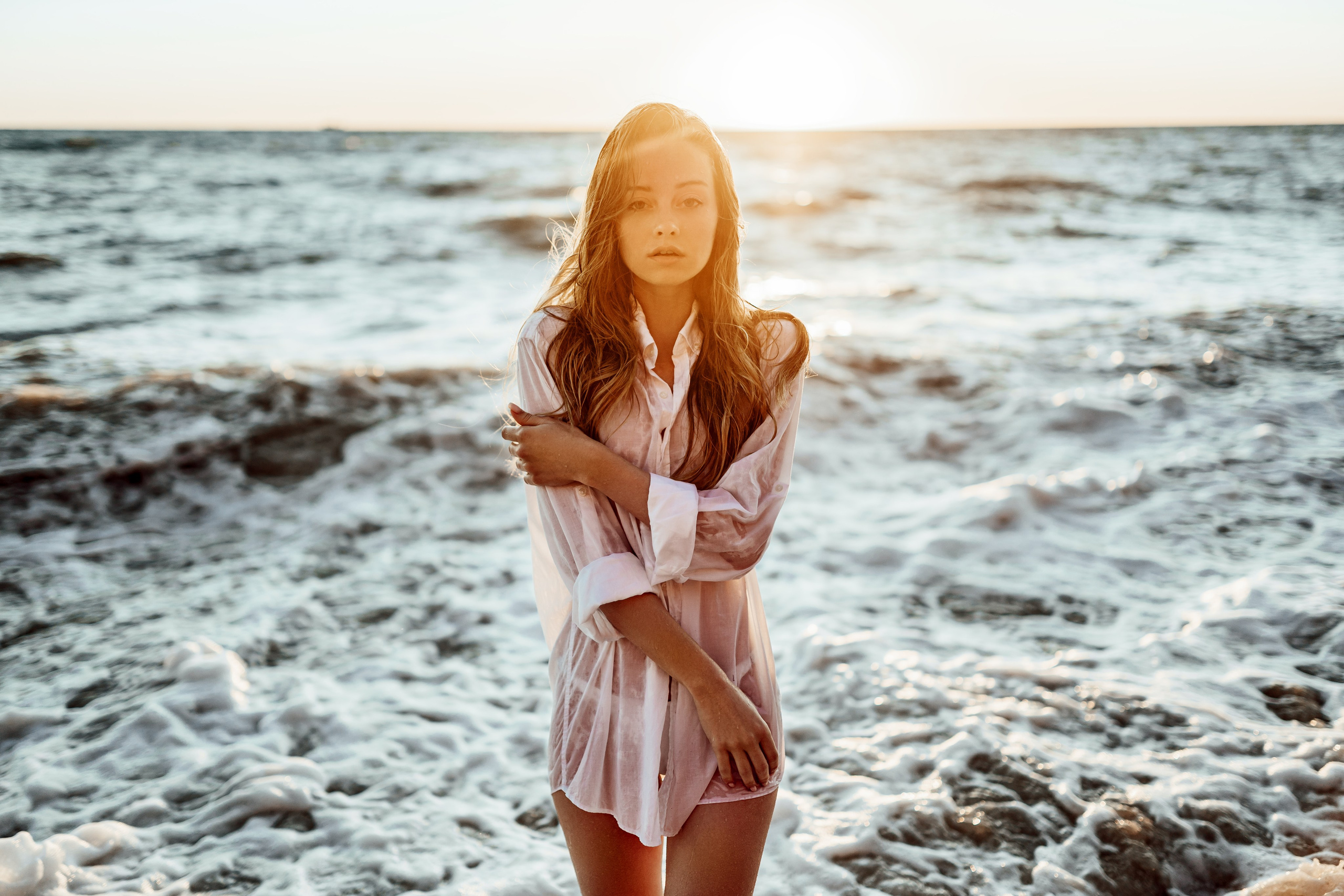 Bathed in the golden hues of sunset, the model stands by the shore in a white bikini and sheer cover-up. The serene ocean and the warm light create a blissful and serene scene.