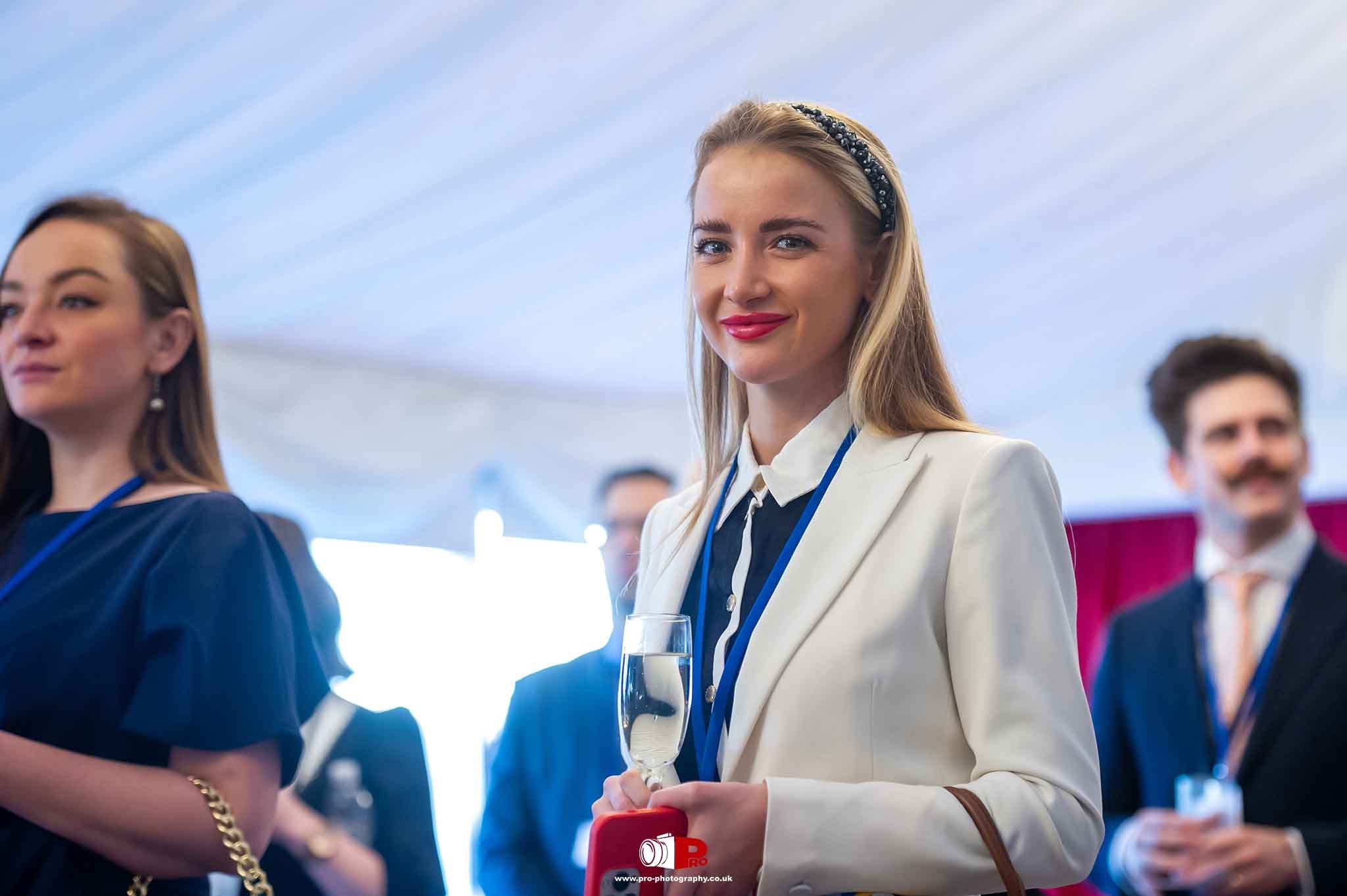 A well-dressed woman in a white blazer holding a champagne glass, smiling at a corporate networking event.