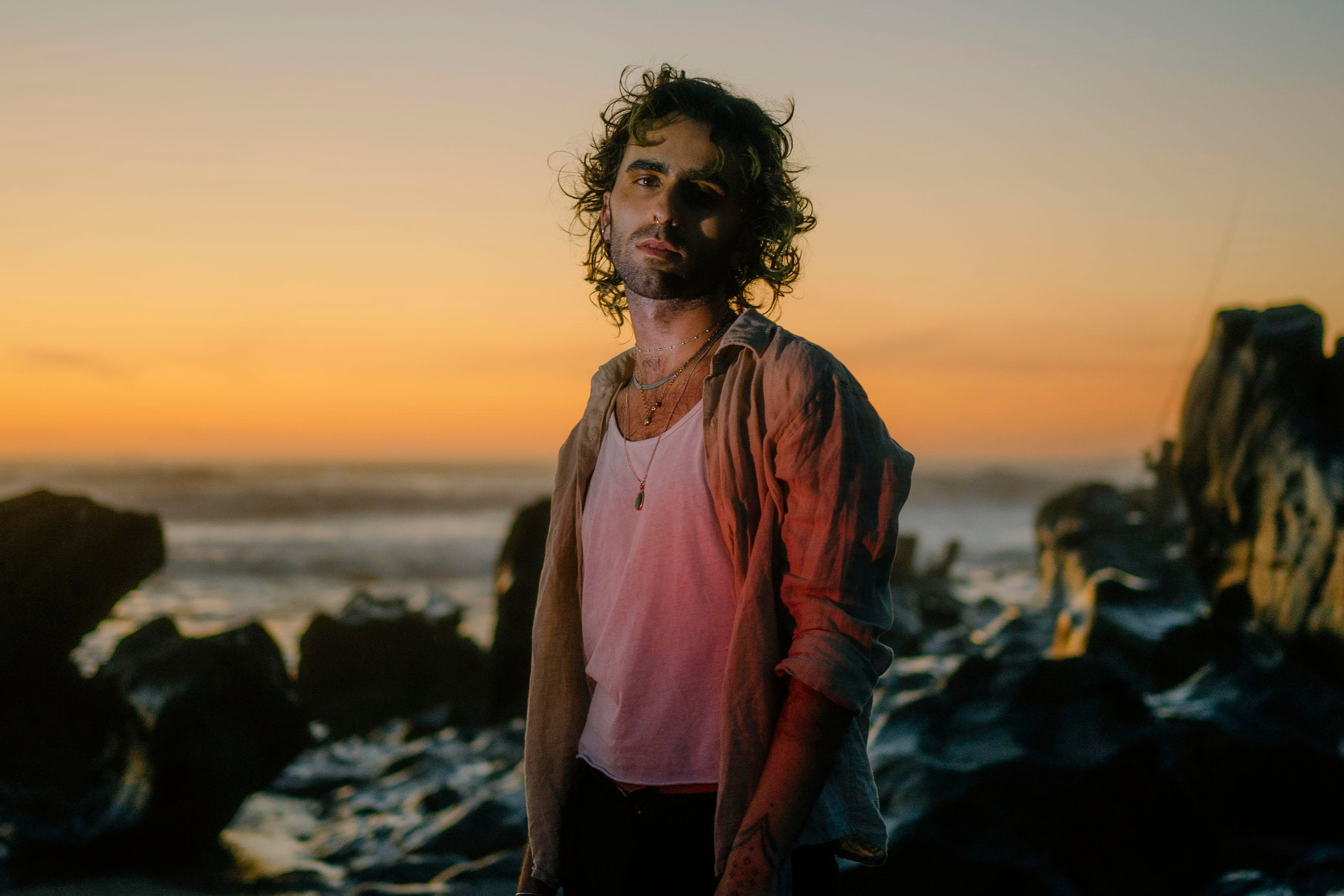 LGBTQ+, LGBT portrait of a beautiful person with makeup, a white tank top, and curly hair. Sunset on a beach, colored light.