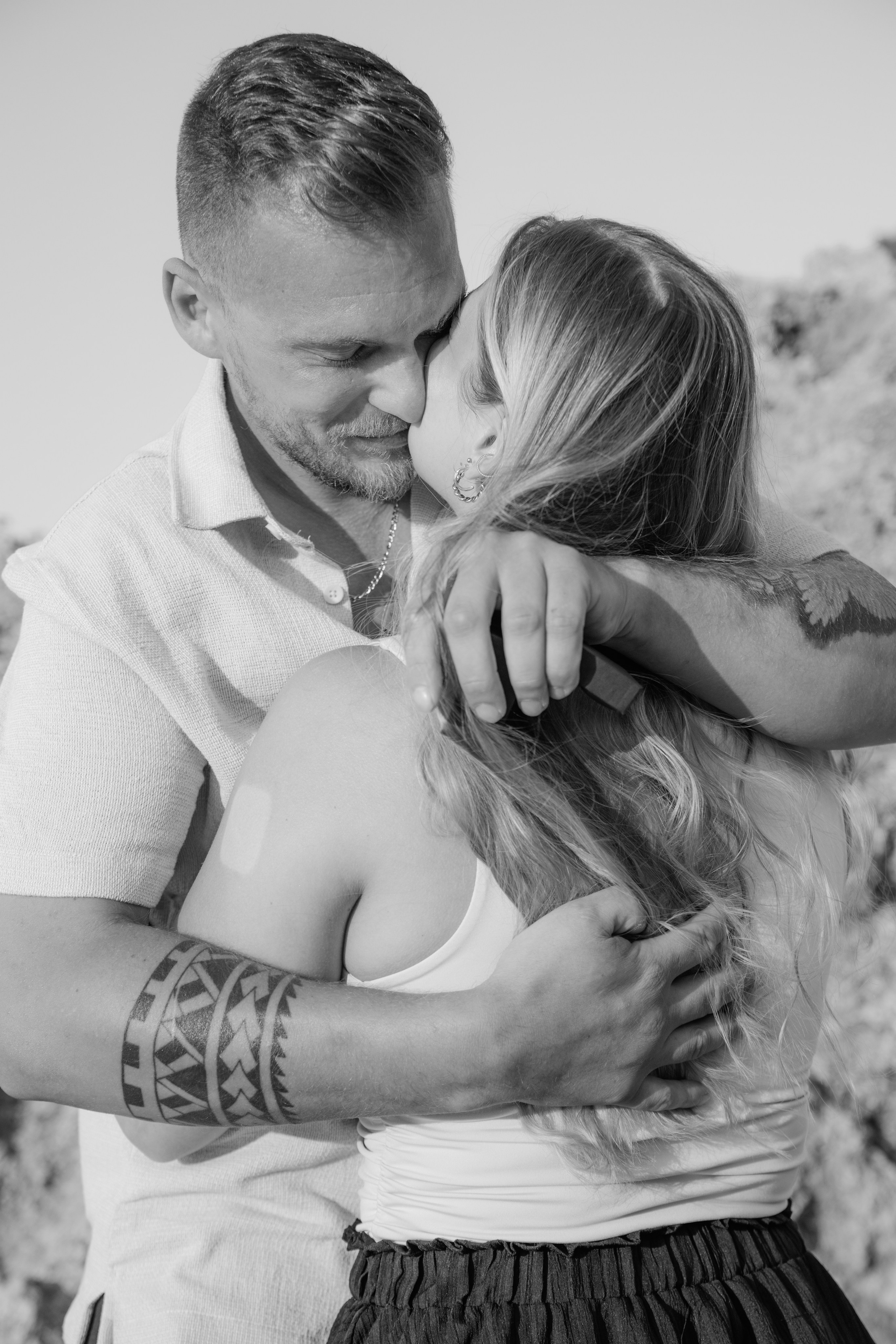 "Innig umarmt am Strand von Kato Petres: Das Paar genießt den ersten Moment nach dem Antrag zwischen den hohen, schützenden Kalksteinfelsen. Die warme Abendsonne beleuchtet die Umarmung, während im Hintergrund die Wellen sanft gegen die steinige Küste von Rhodos schlagen."