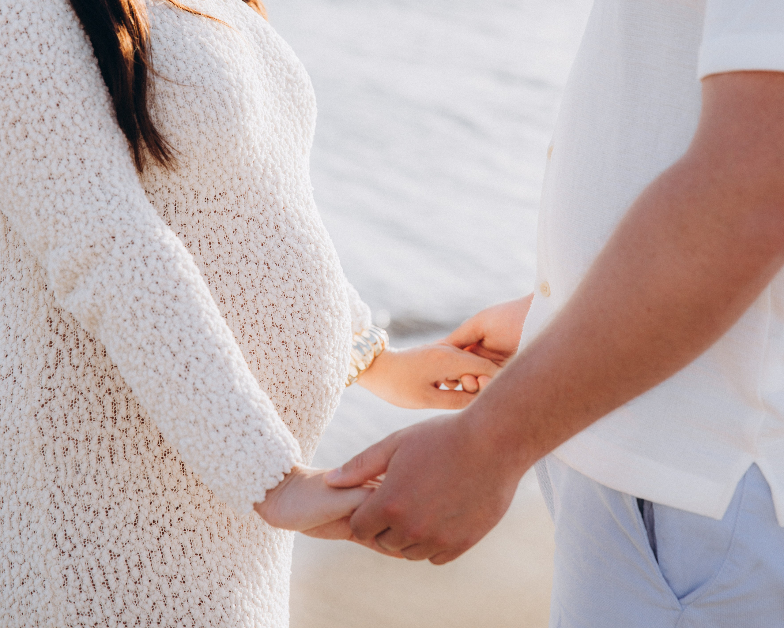 Tierno momento de embarazo junto al mar en Jávea, España — primer plano de una pareja tomándose de las manos cerca del vientre. Perfecto para quienes buscan sesiones íntimas de maternidad en Jávea y la costa española.