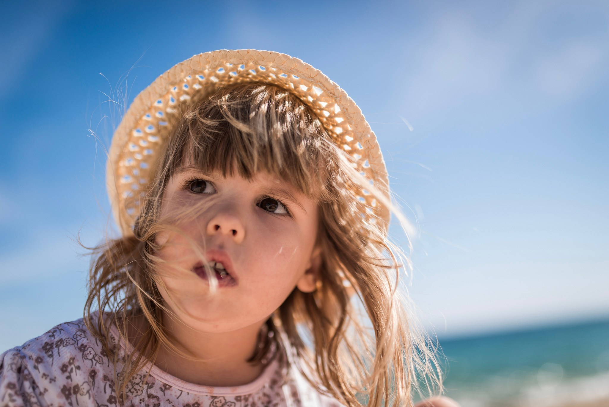 Krupni kadar iz niskog ugla devojčice u slamnom šeširu, sa kosom koju vetar raznosi, koja gleda ka vedrom plavom nebu tokom sunčanog dana na plaži.