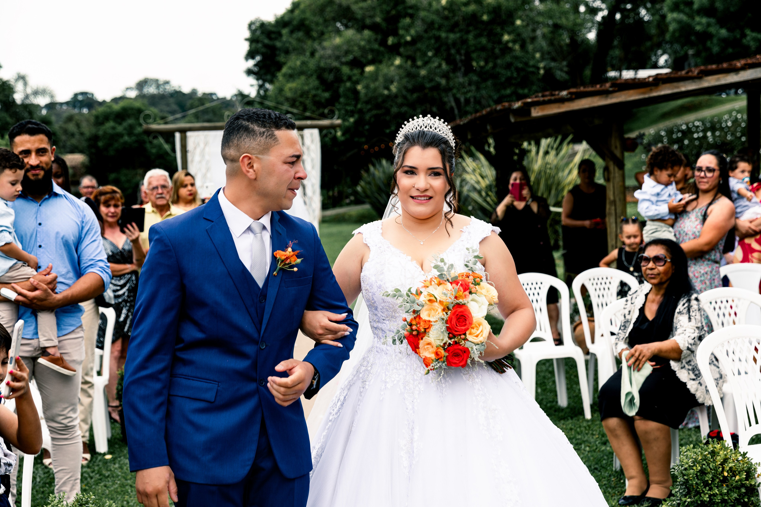 Casal sorrindo em sua entrada na cerimônia, com padrinhos e convidados celebrando o momento.