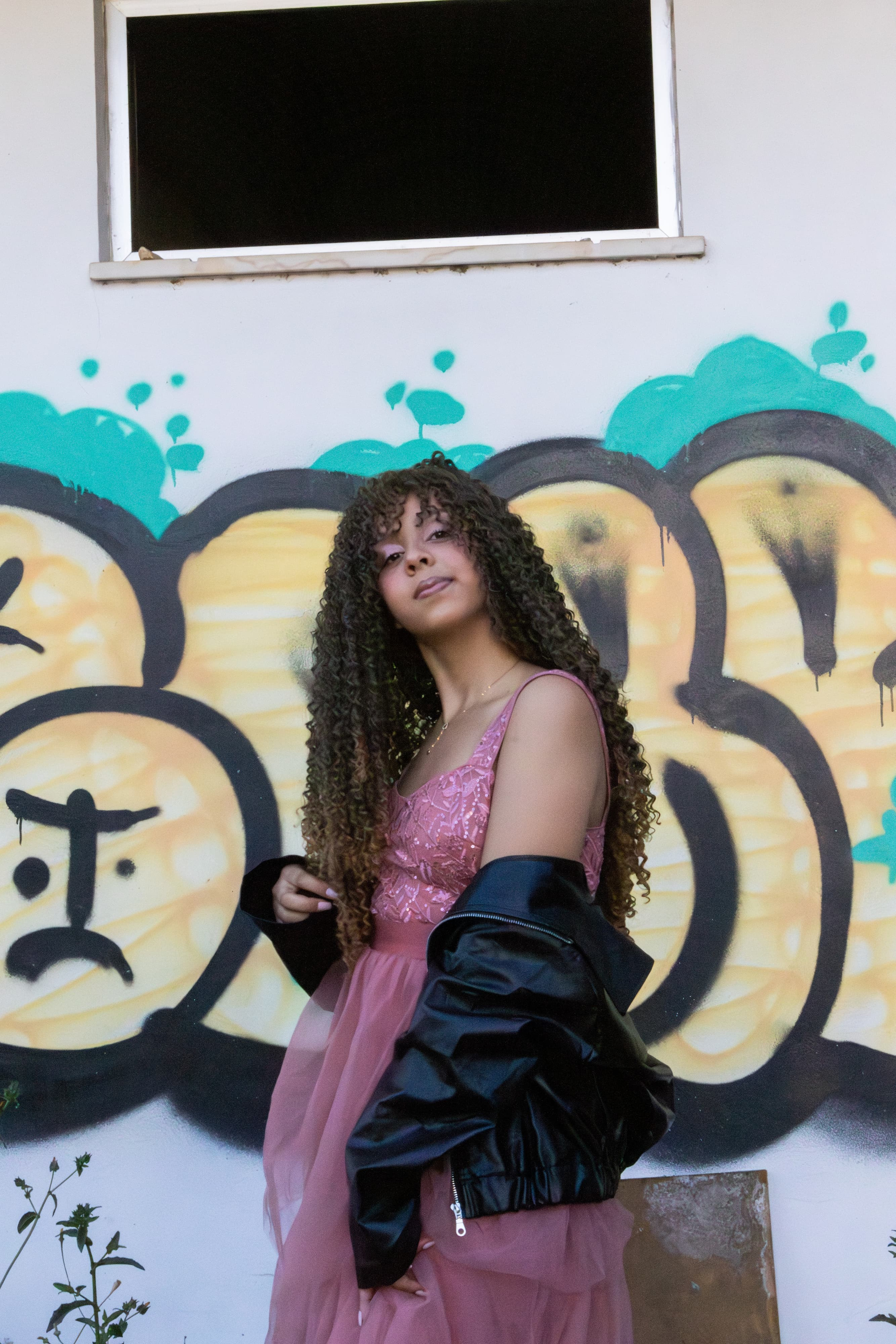 Jovem sorrindo com jaqueta de couro preta e vestido rosa, posando em frente a um muro urbano com grafites coloridos. Estética urbana e delicada.
