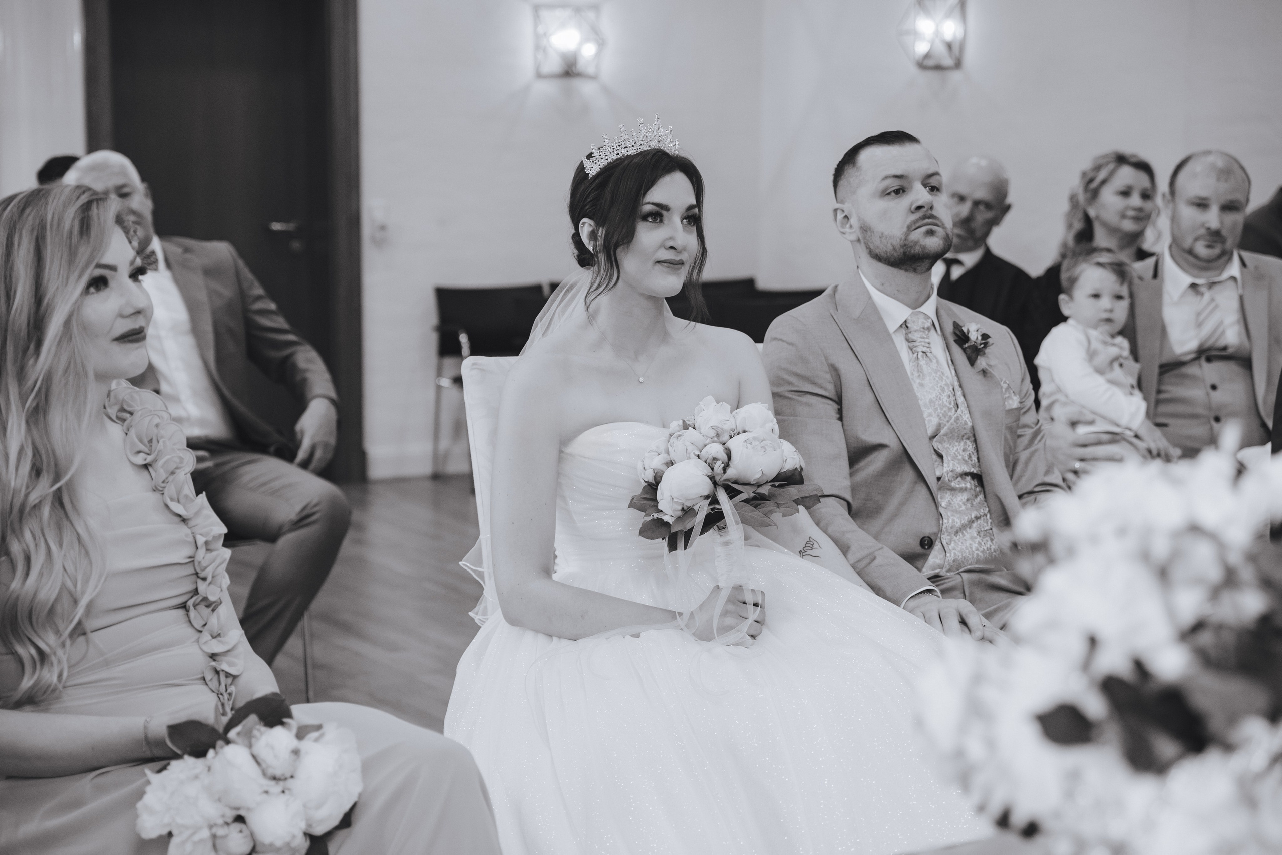 Schwarz-Weiß-Foto, auf dem der Bräutigam, die Braut und die Gäste der Rede der Standesbeamtin während einer Hochzeit im Standesamt zuhören.