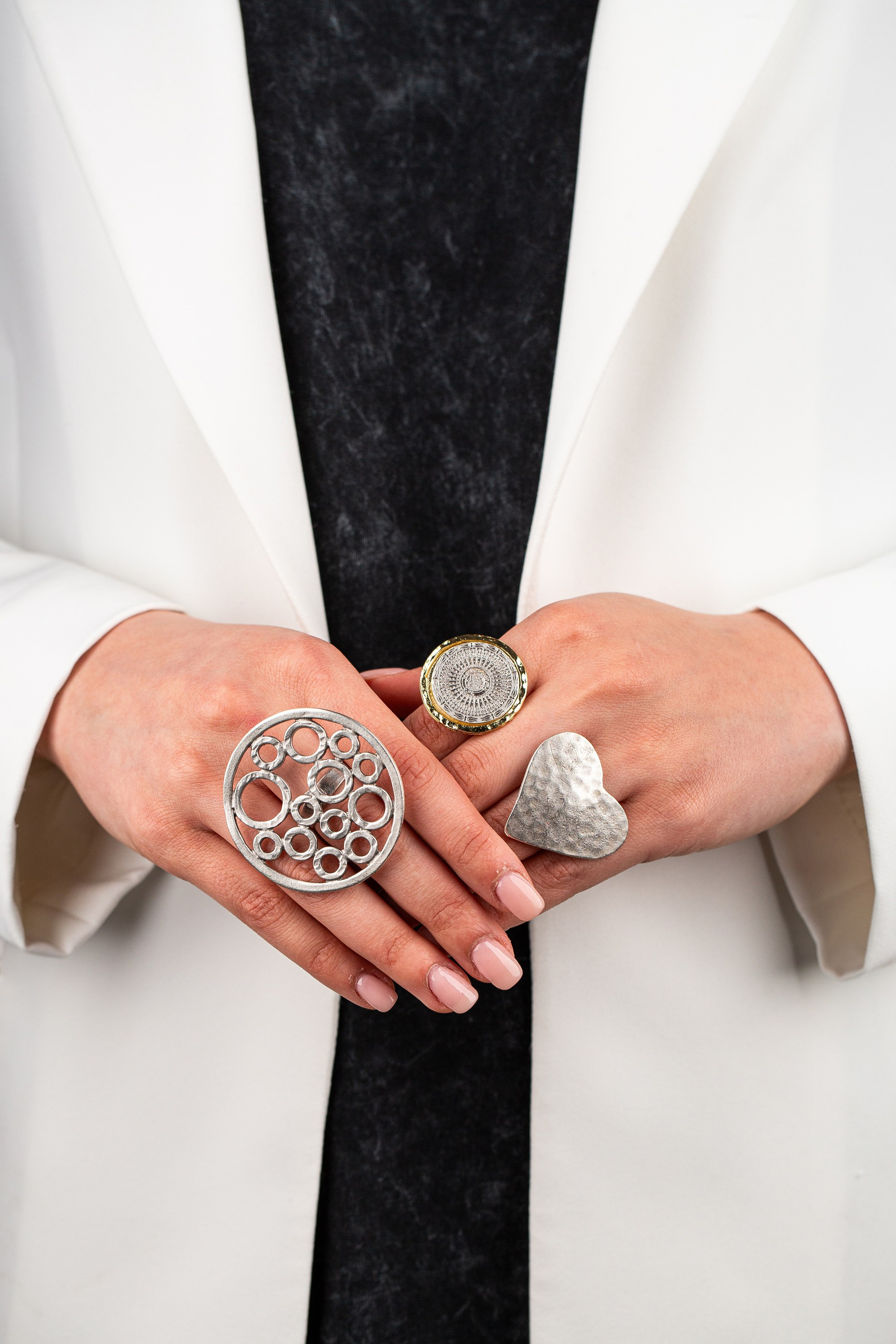 Gümüş və qızılı tonlarda həndəsi və ürək formalı iri üzüklər taxmış ağ gödəkçəli şəxsin əlləri.