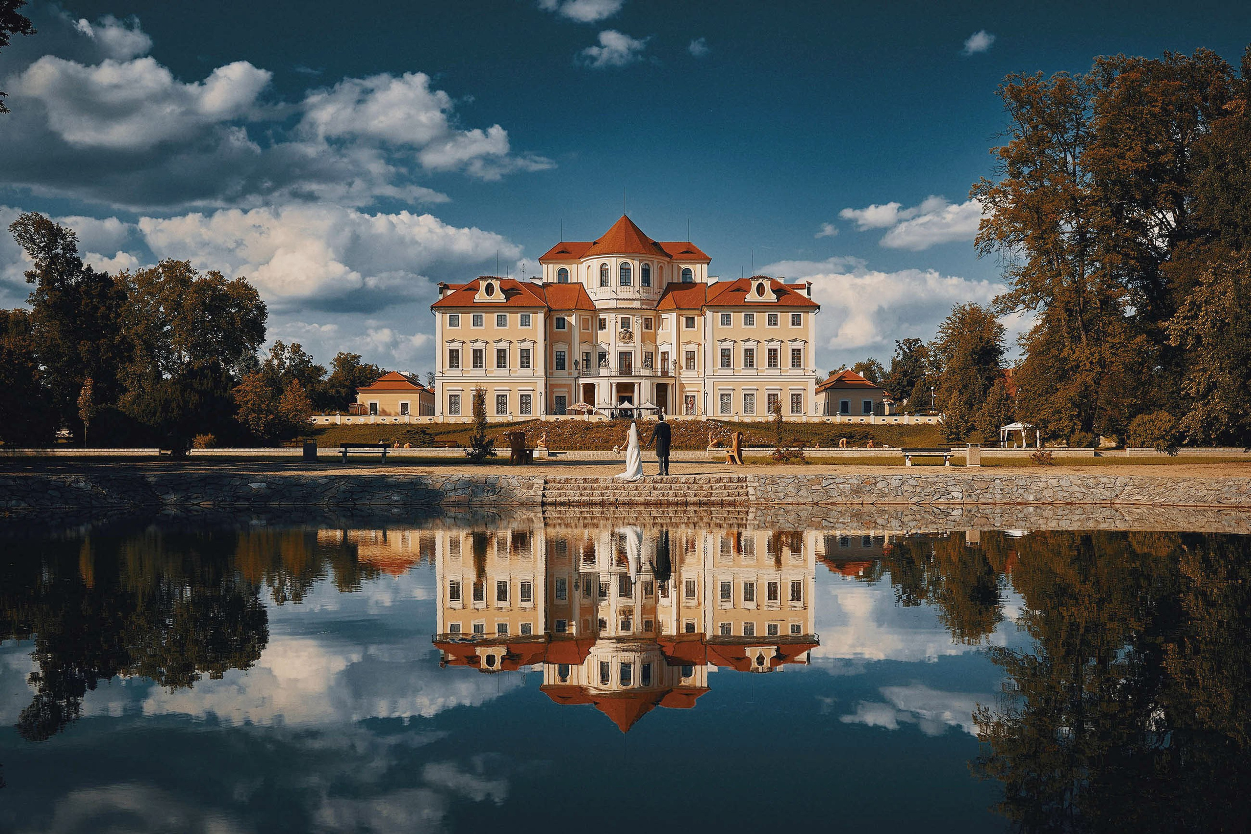 Couple facing the historic chateau across a reflective lake, with vibrant blue skies mirrored in the water.