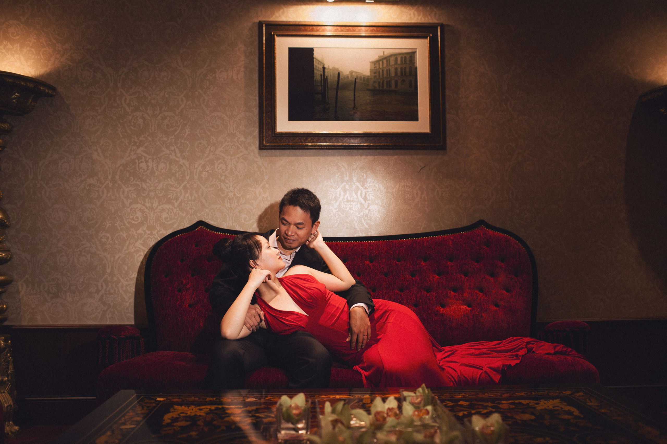 Thai bride-to-be in red dress lying in fiance's lap embraced inside Hotel Danieli Venice.