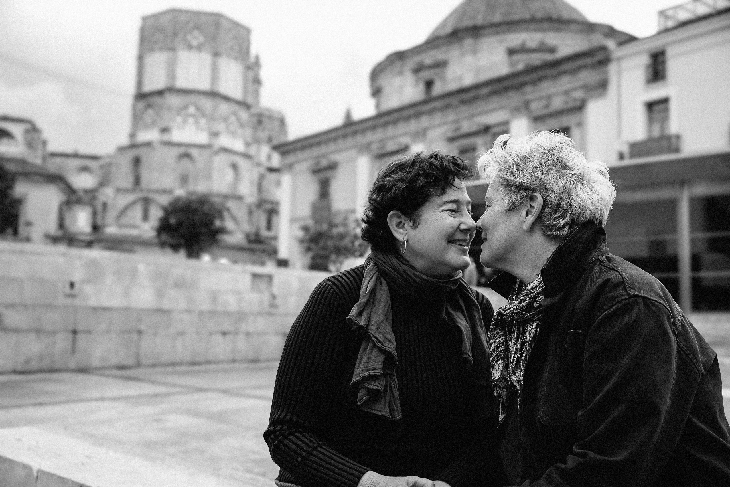 Семейная фотосессия в Валенсии, Испания, где две женщины делят близкий эмоциональный момент на исторической городской площади, снято в чёрно-белом стиле для вневременной и интимной семейной связи — идеально для семейных фотосессий в Валенсии и по всей Испании.