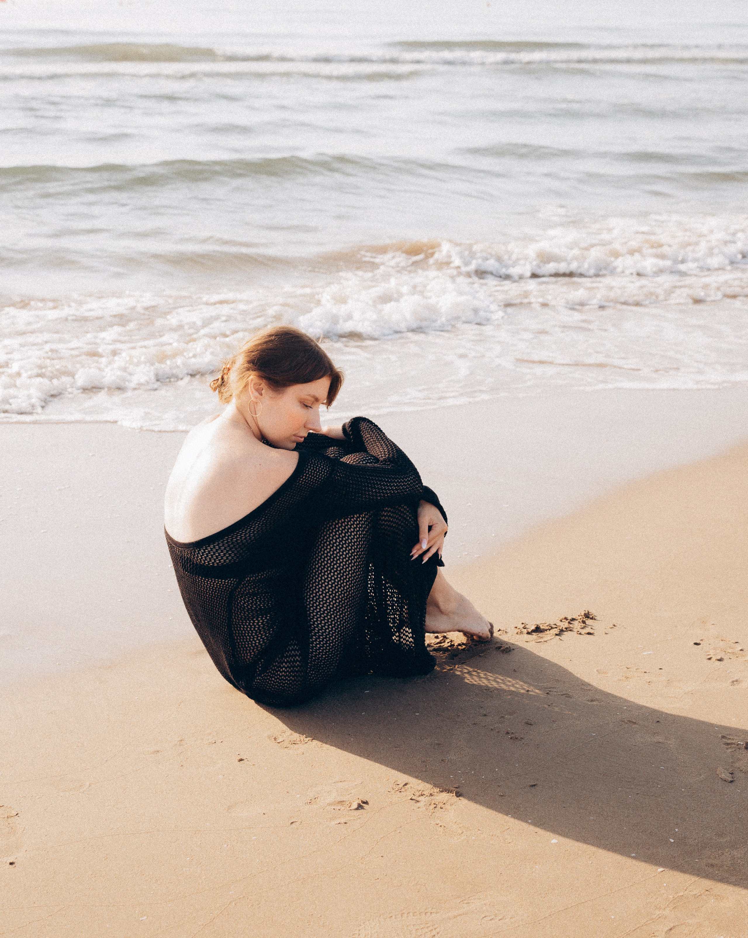 Женщина в полупрозрачном черном платье сидит на песке на закате, задумчиво смотрит вниз, пока волны мягко касаются берега в Кальпе, Испания — эмоциональная и художественная пляжная портретная фотография, передающая спокойствие и природную красоту, идеально подходит для портретных фотосессий в Кальпе и Испании.