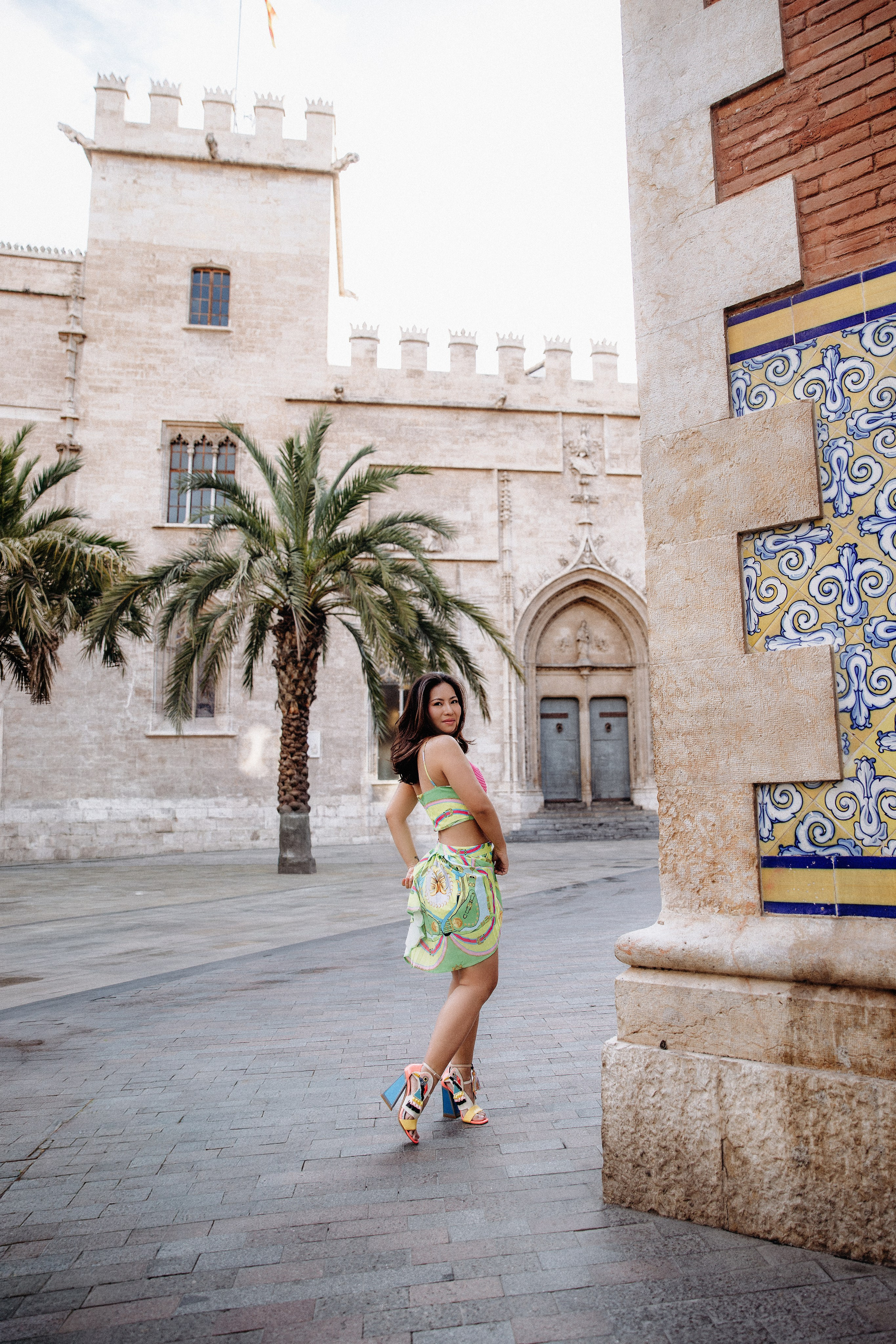 Sesión de retrato femenino en Valencia, España, con una mujer vestida con un elegante vestido caminando por una histórica plaza rodeada de palmeras y arquitectura mediterránea clásica. Capturada con luz natural para crear un retrato sofisticado, inspirado en el estilo de vida y atemporal, ideal para fotografía de retrato femenino, retratos de moda y sesiones individuales en Valencia y toda España.