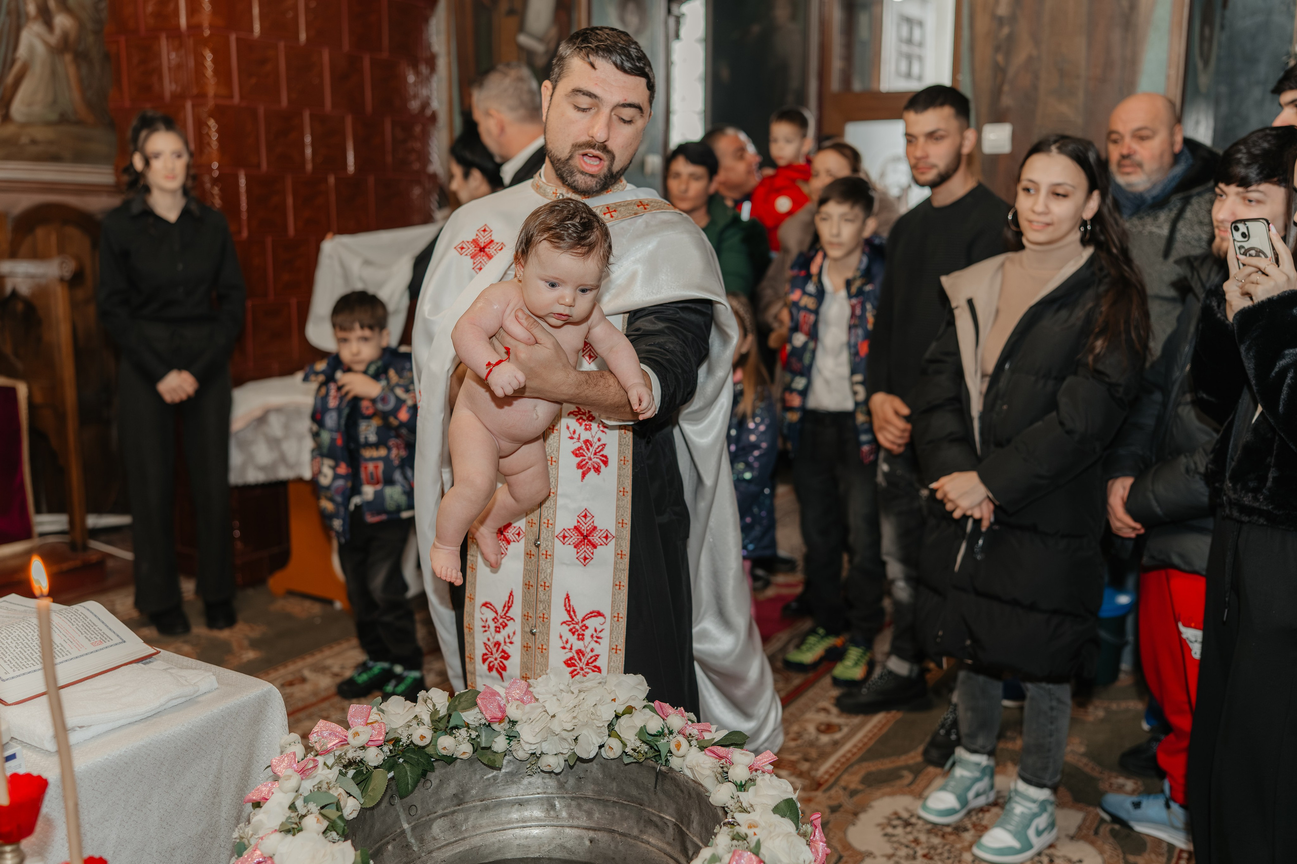 Momentul imediat înaintea scăldării: preotul ține pruncul Eveline Maria în brațe, gata să-l introducă în cristelniță pentru botez. O fotografie aanticipației și a solemnității ritualului sfânt.