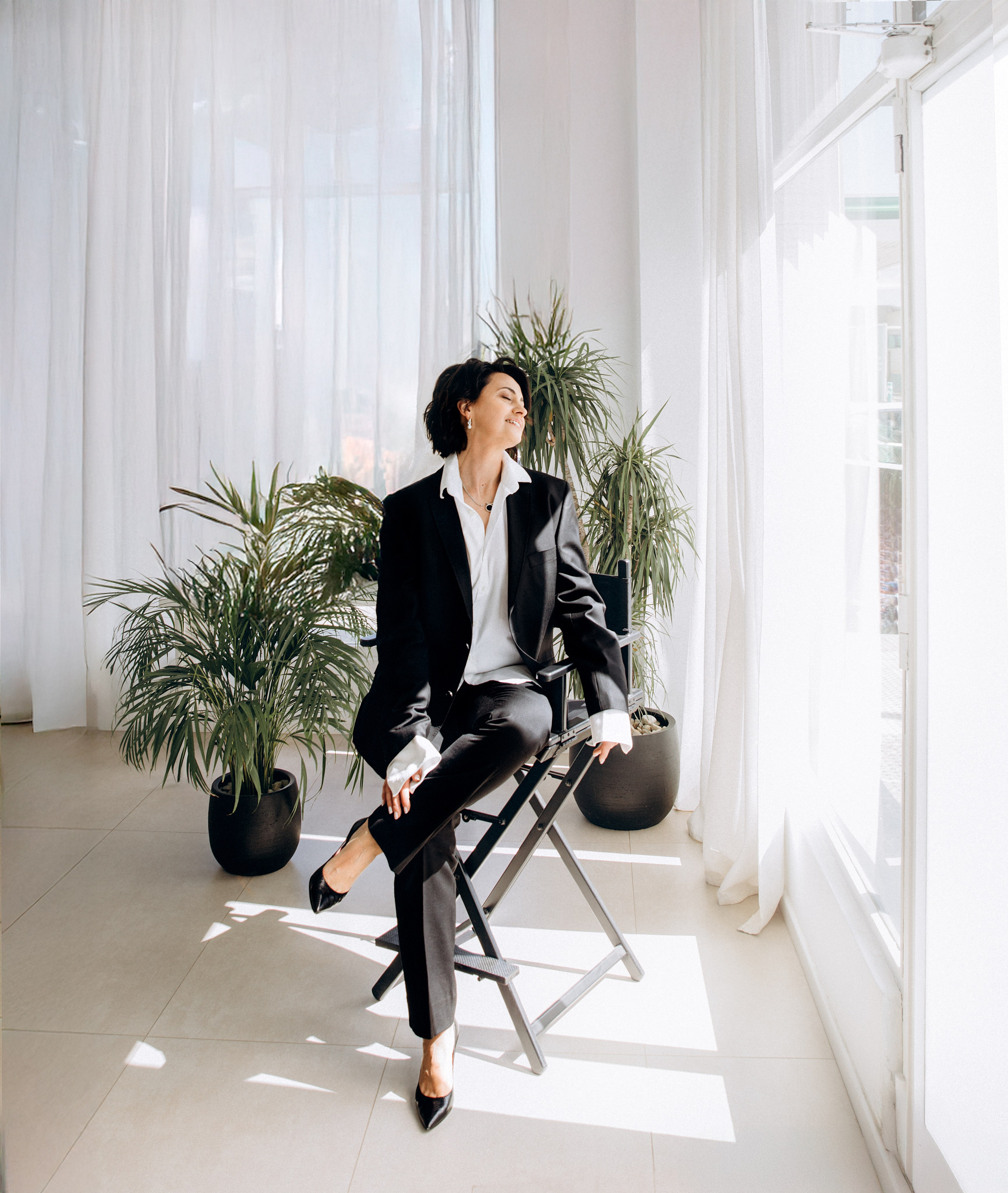 Стильная женщина в черном костюме и на каблуках, сидящая в современной фотостудии с тропическими растениями и белыми шторами, в Валенсии, Испания