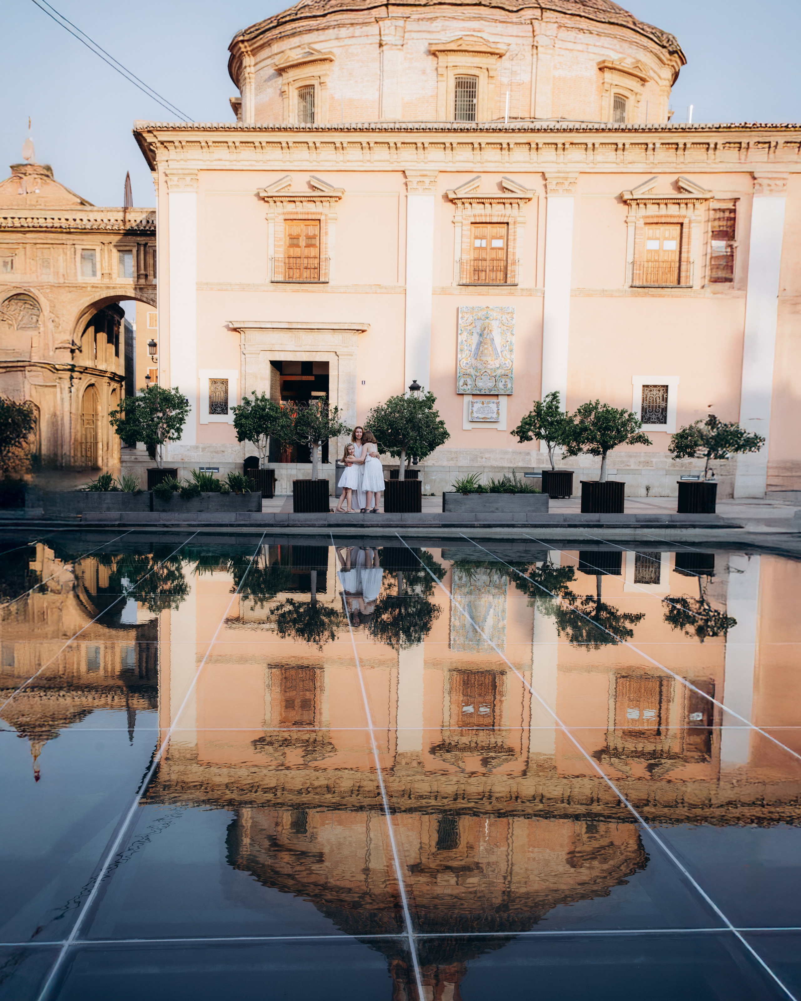 Madre e hijas vestidas de blanco posan frente a la histórica Basílica de la Virgen de los Desamparados en Valencia, España, con su reflejo en un estanque cercano. Impresionante sesión familiar en Valencia que captura arquitectura atemporal y conexión emocional, ideal para quienes buscan fotografía familiar elegante en España.