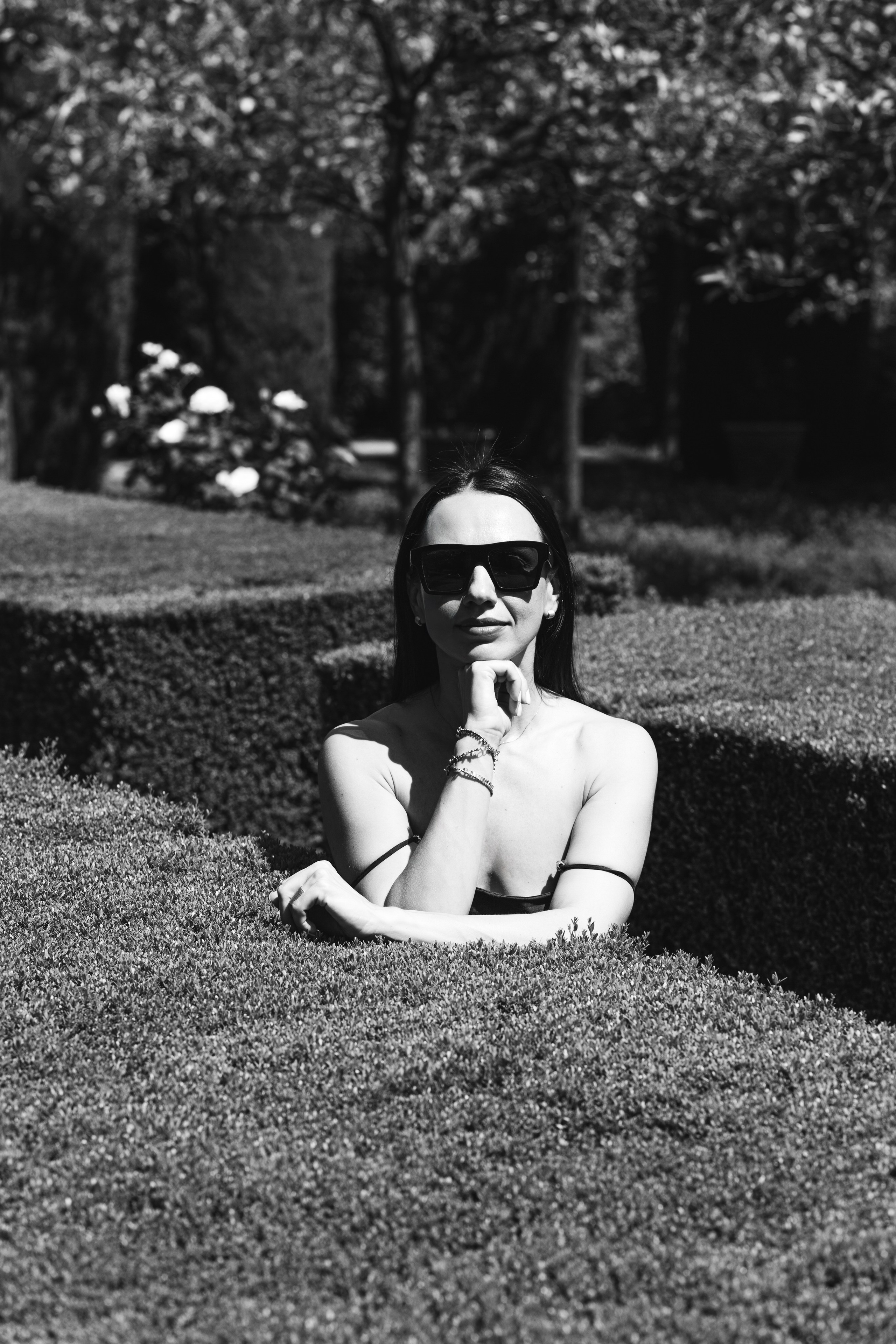 Стильная женщина в солнцезащитных очках с расслабленным выражением лица позирует на фоне фигурных кустов в саду в Дении, Испания — черно-белая художественная портретная фотография, подчеркивающая элегантность, контраст и природное окружение, идеально подходит для портретных фотосессий в Дении и Испании.