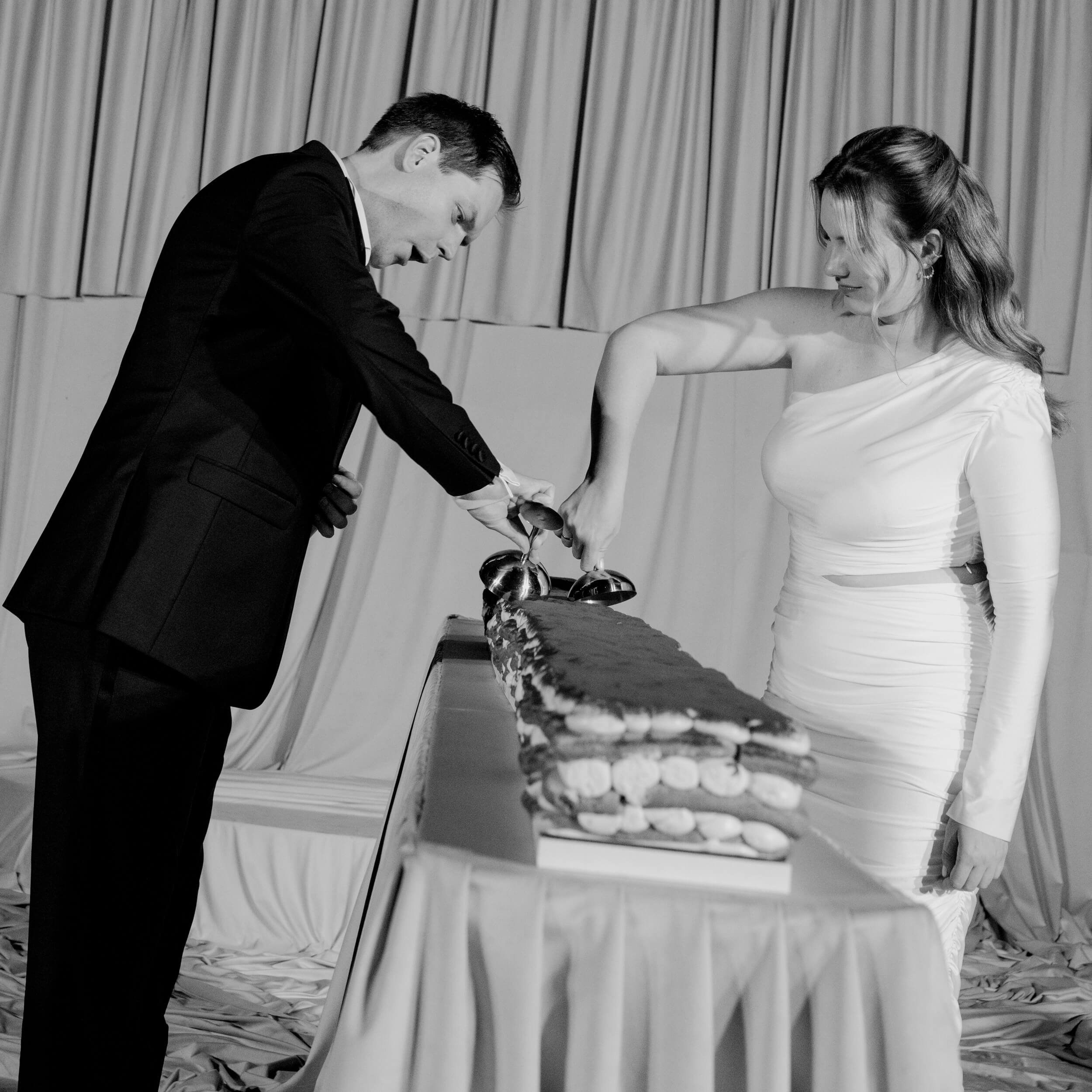Black and white photo of the groom bowing gracefully toward the bride in a white satin gown in front of a draped curtain backdrop, playful couple moment at Lake Como wedding