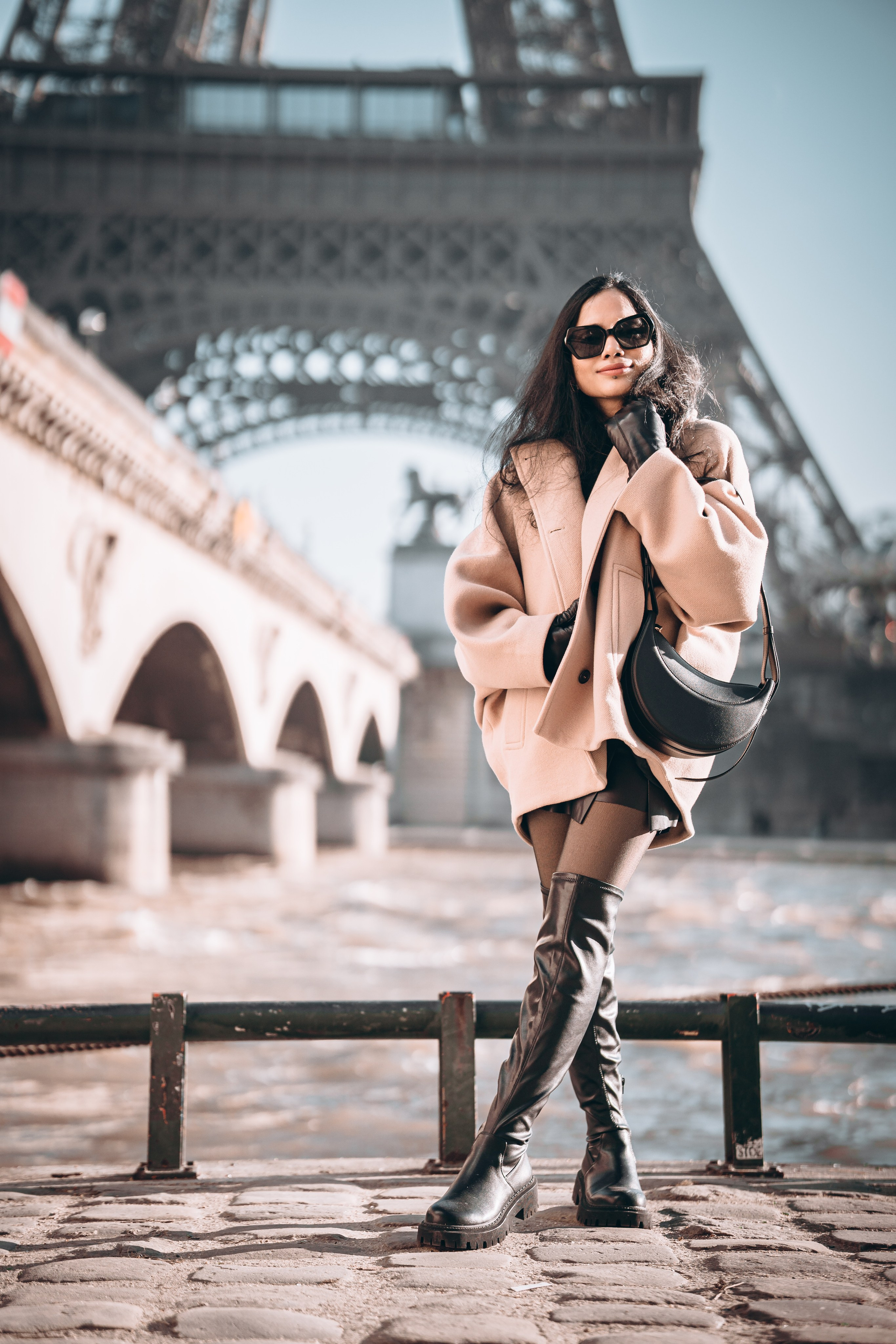 a woman wearing oversized brown jacket and standing with paris bridge and eiffel tower view behind her