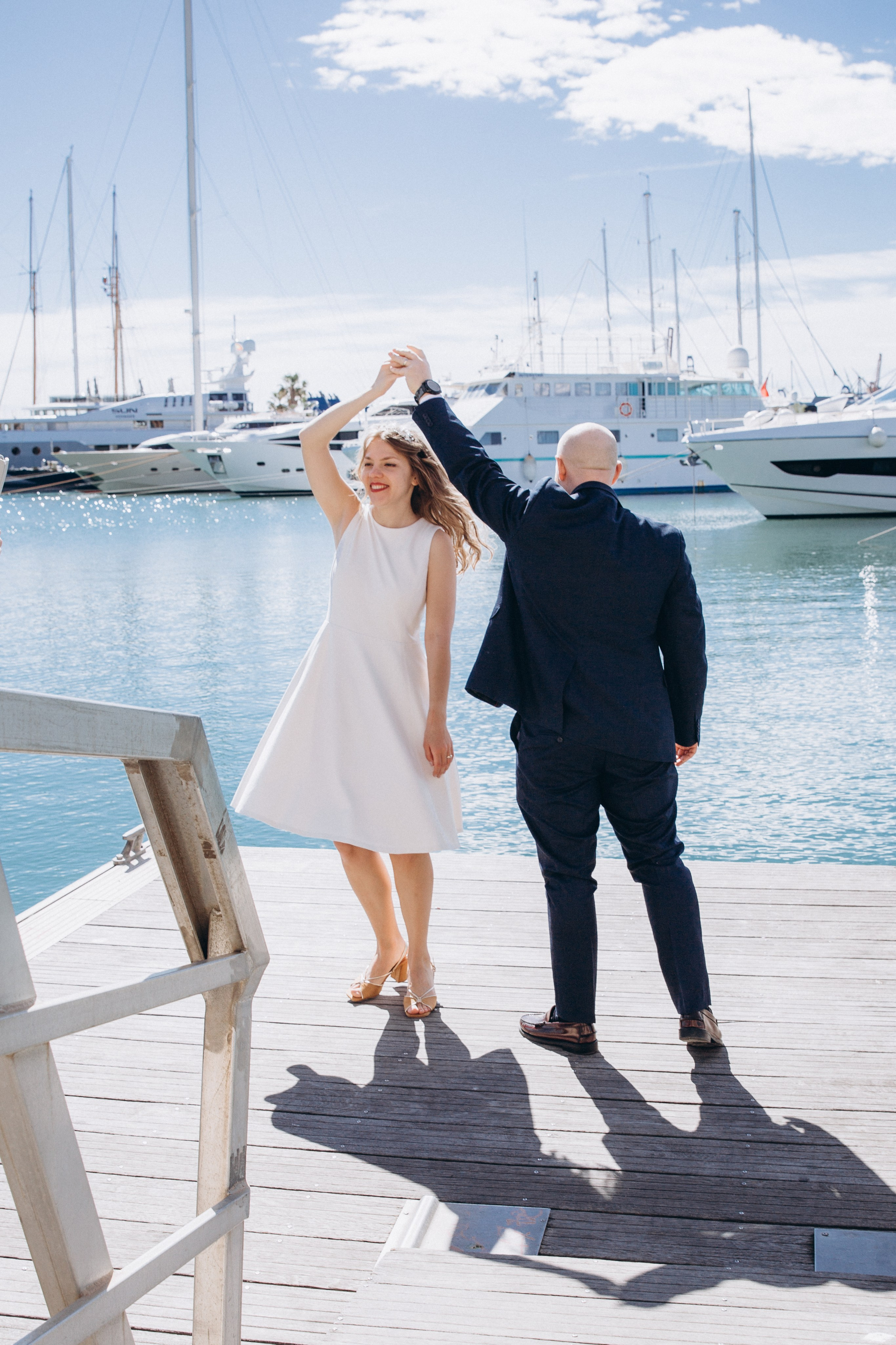 Romantic wedding dance on a marina dock in Valencia, Spain — joyful bride twirling in a white dress as the groom lifts her hand, with luxury yachts and blue skies in the background. Perfect for couples seeking elegant and spontaneous wedding photoshoots in Valencia and along Spain’s Mediterranean coast.