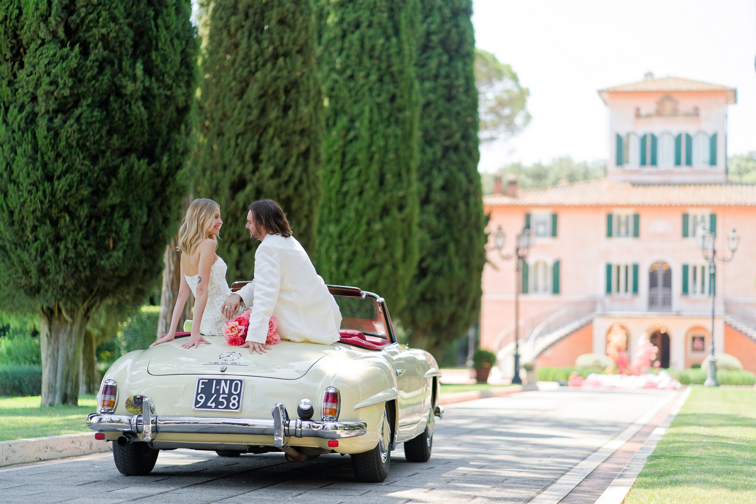 "Newlyweds driving a white vintage convertible on a cypress-lined road near an Italian villa."