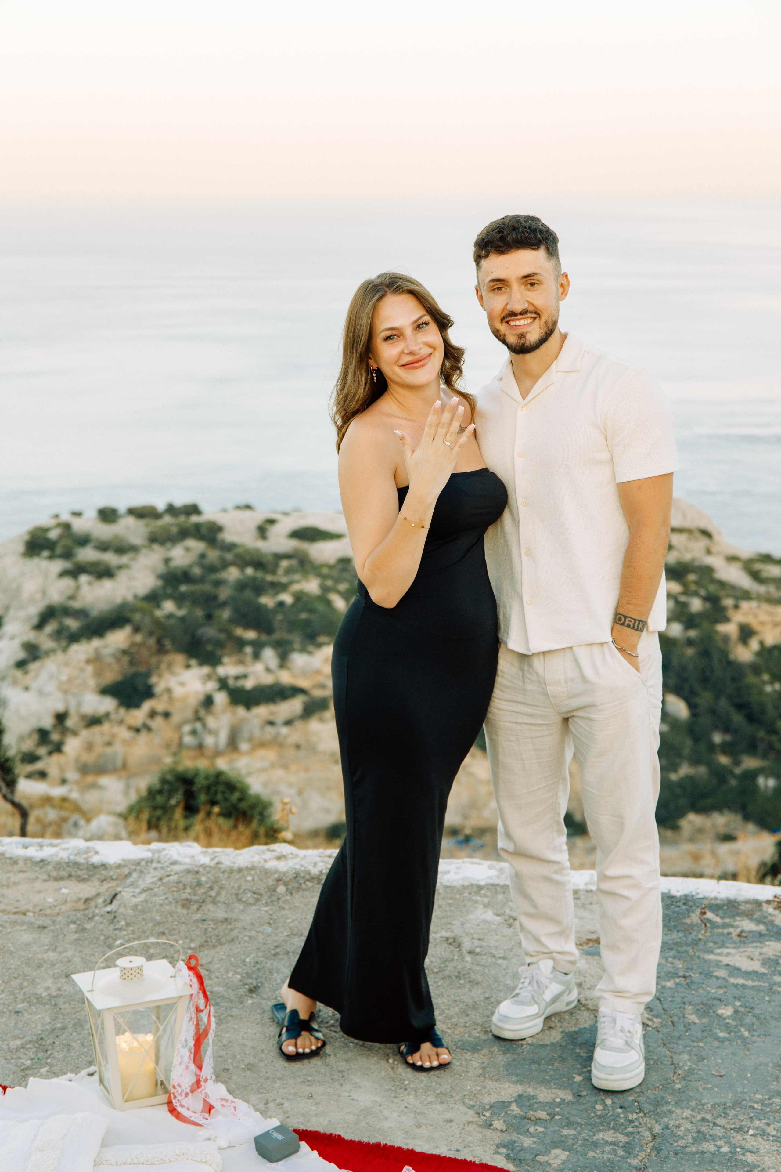 "Ein glückliches, frisch verlobtes Paar beim Fotoshooting auf dem Gipfel des Profitis Ilias. Sie stehen eng umschlungen auf einem Felsplateau, lachen in die Kamera und zeigen stolz den Verlobungsring. Im Hintergrund erstreckt sich das beeindruckende Panorama der Bucht von Faliraki und das glitzernde Ägäische Meer."