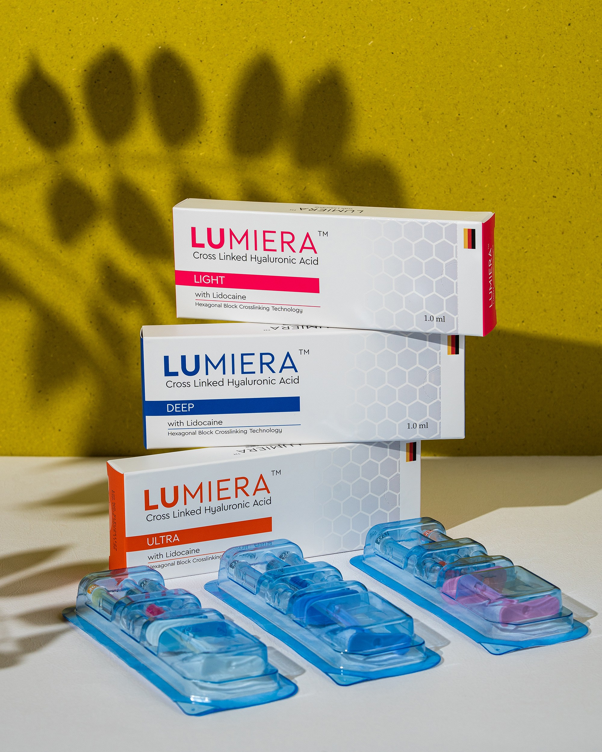 Three types of LUMIERA hyaluronic acid fillers (Light, Deep, and Ultra) stacked against a yellow background with leaf shadows, with corresponding syringes in blue trays in the foreground.