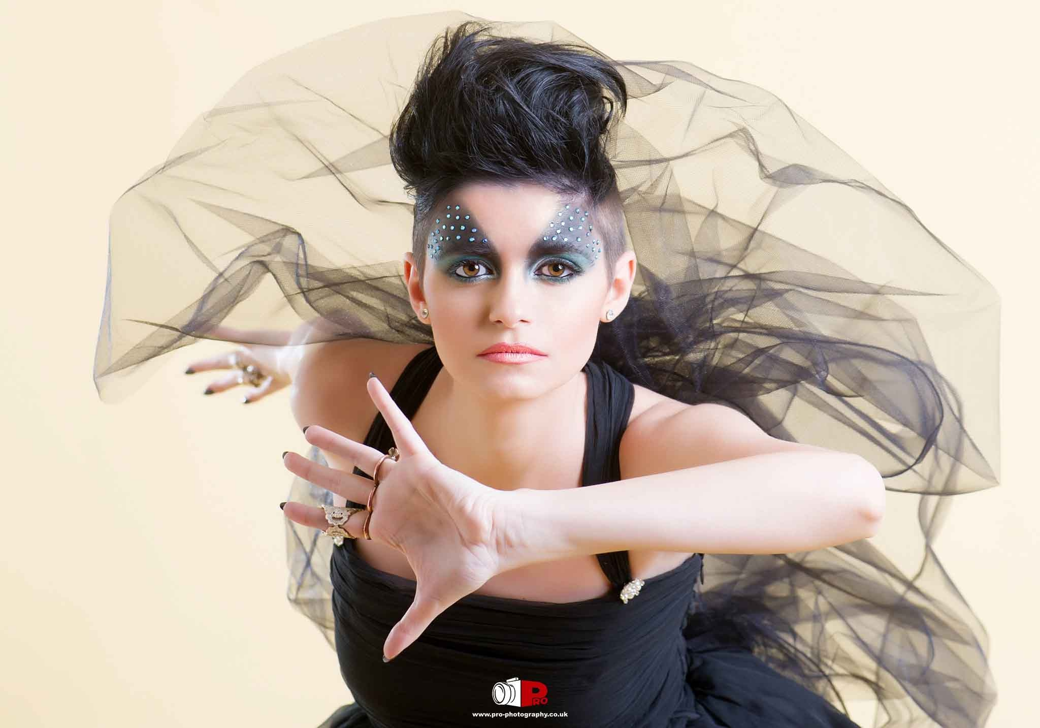 Artistic fashion portrait of a woman with striking eye makeup, embellished with crystals, and a voluminous black tulle dress, posing with expressive hand gestures.