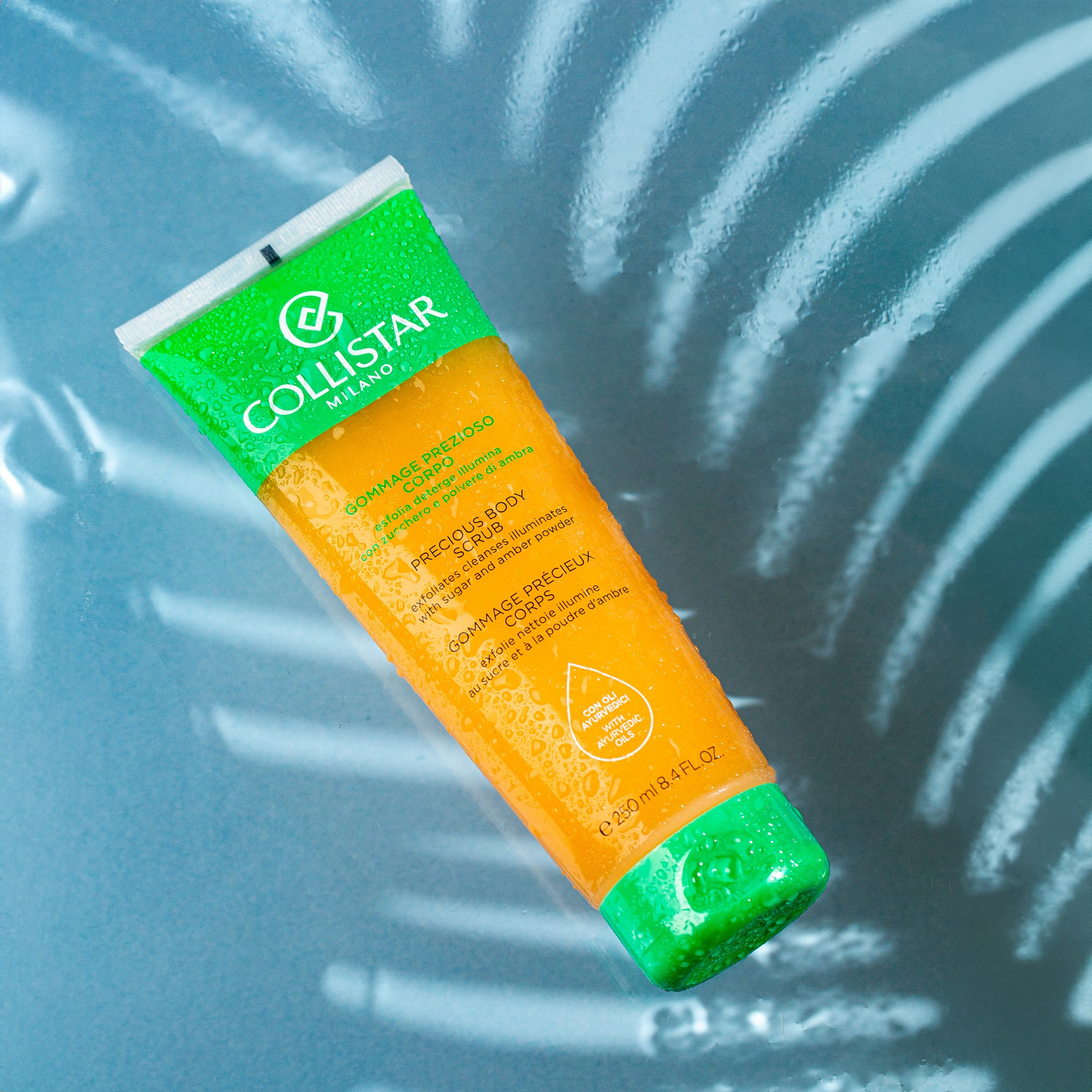 Collistar Precious Body Scrub, su damcıları ilə örtülmüş, mavi fon və işıq naxışları ilə təqdimat.