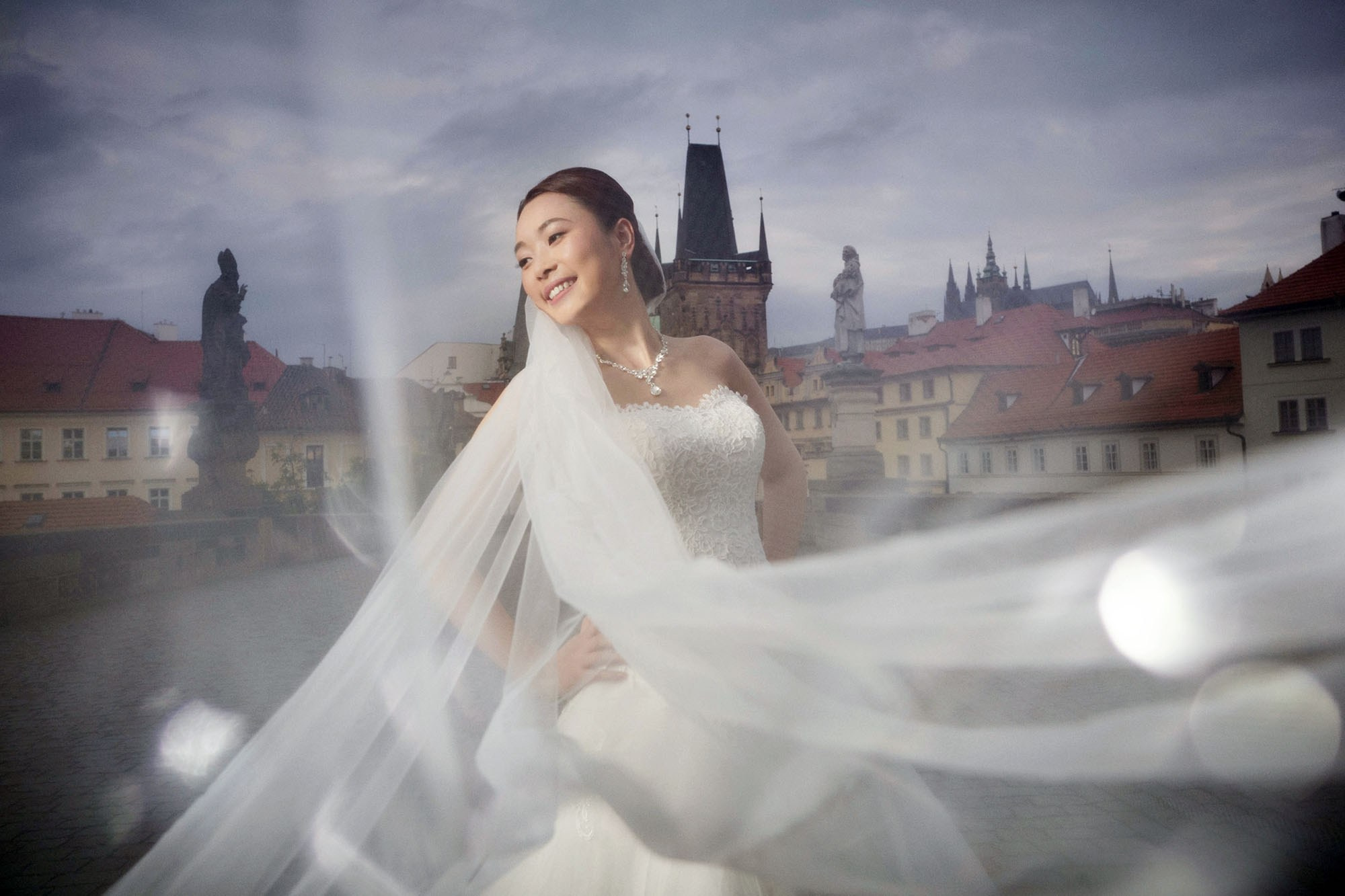 Joyful Chinese bride surrounded by her wedding veil on the historic Charles Bridge at dusk in Prague.