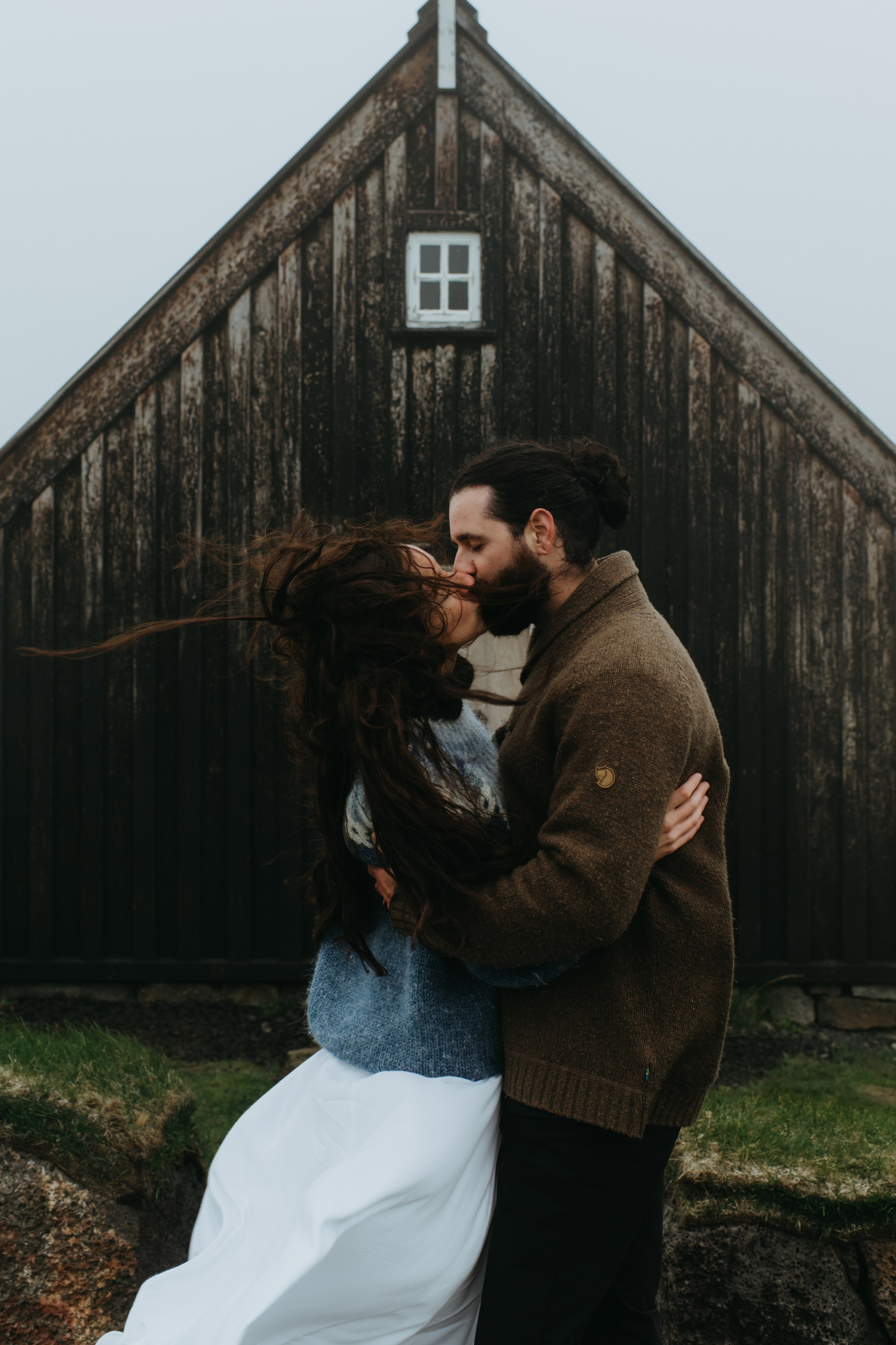 Bride in a flowing dress and groom in a traditional Icelandic sweater, walking across the rocky terrain of Krýsuvík.