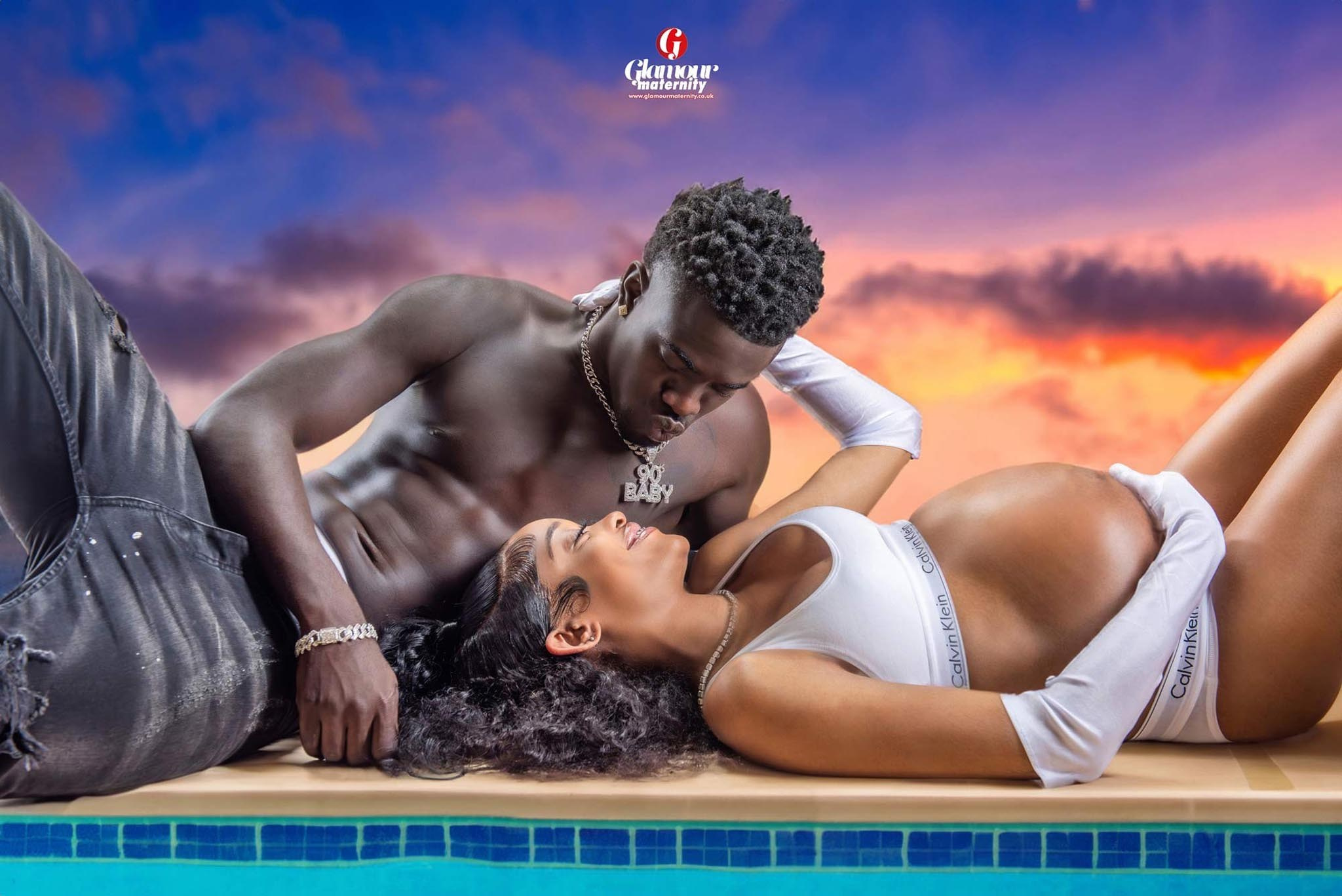 Modern maternity portrait of an expectant couple reclining together in white styling with a warm sunset background.