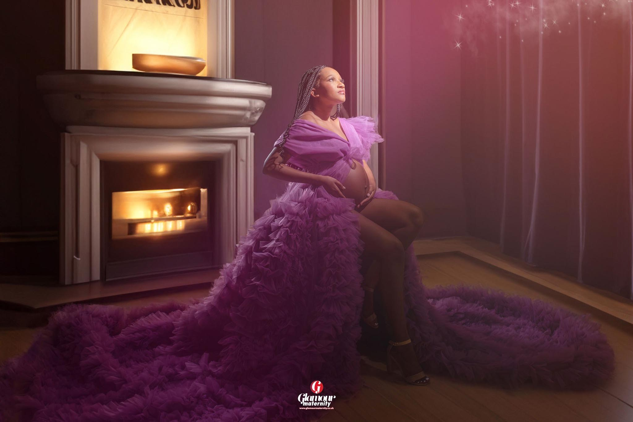 Elegant maternity portrait in a purple ruffled gown beside a glowing fireplace with soft romantic lighting.
