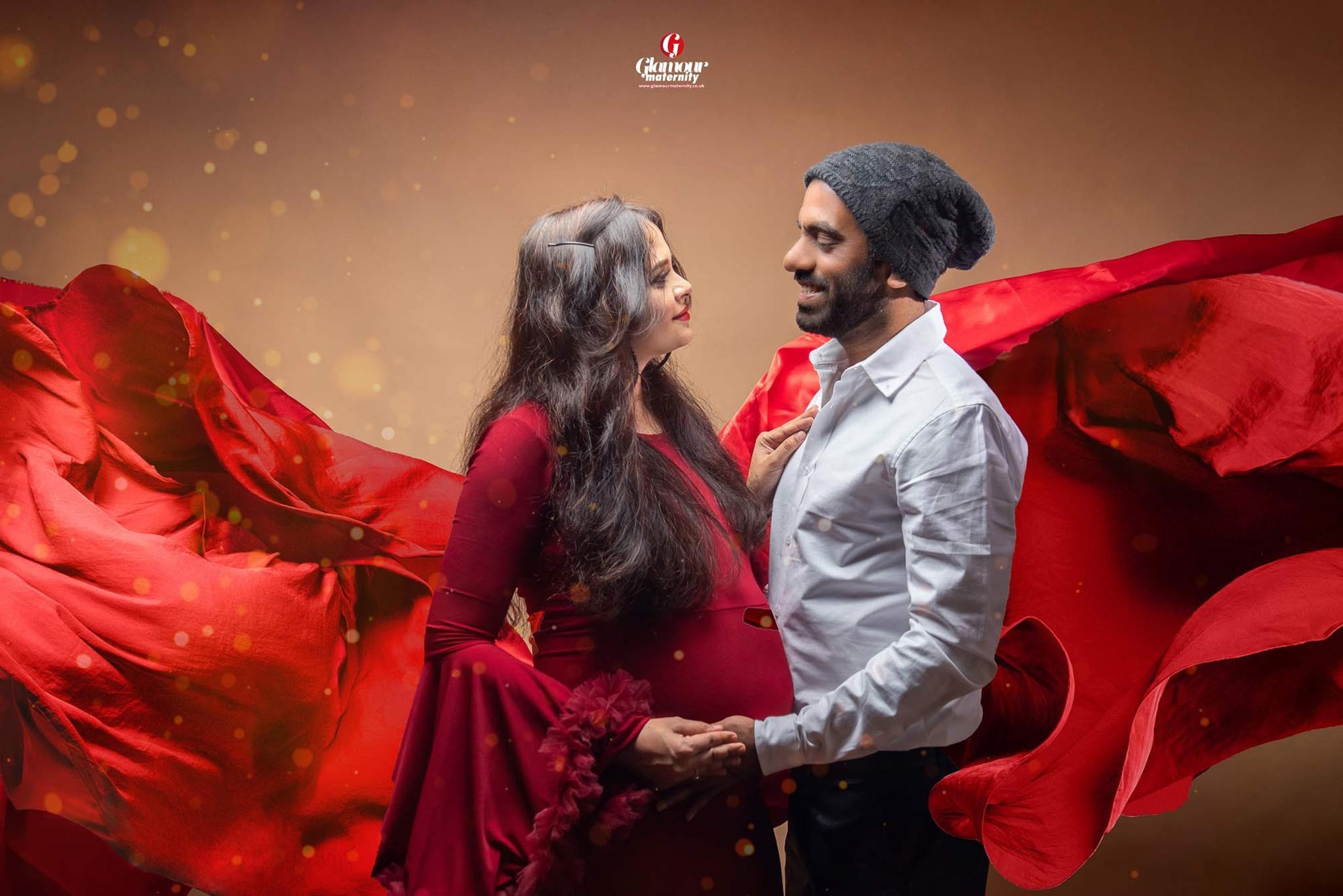 Glamorous maternity portrait of a couple surrounded by dramatic flowing red fabric and warm studio lighting.