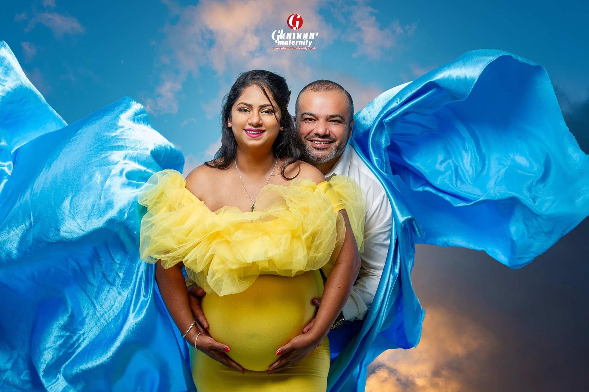 Bright maternity portrait of an expectant couple in yellow and blue styling with dramatic flowing fabric.