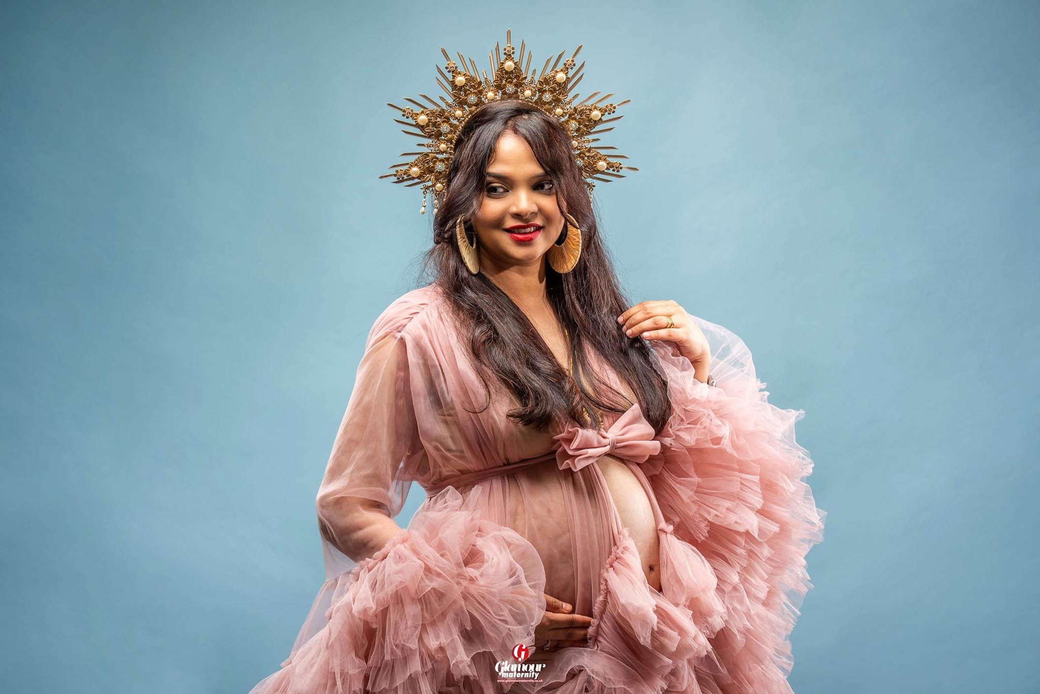 Glamour maternity portrait featuring a soft pink ruffled gown, ornate gold crown and blue studio background.