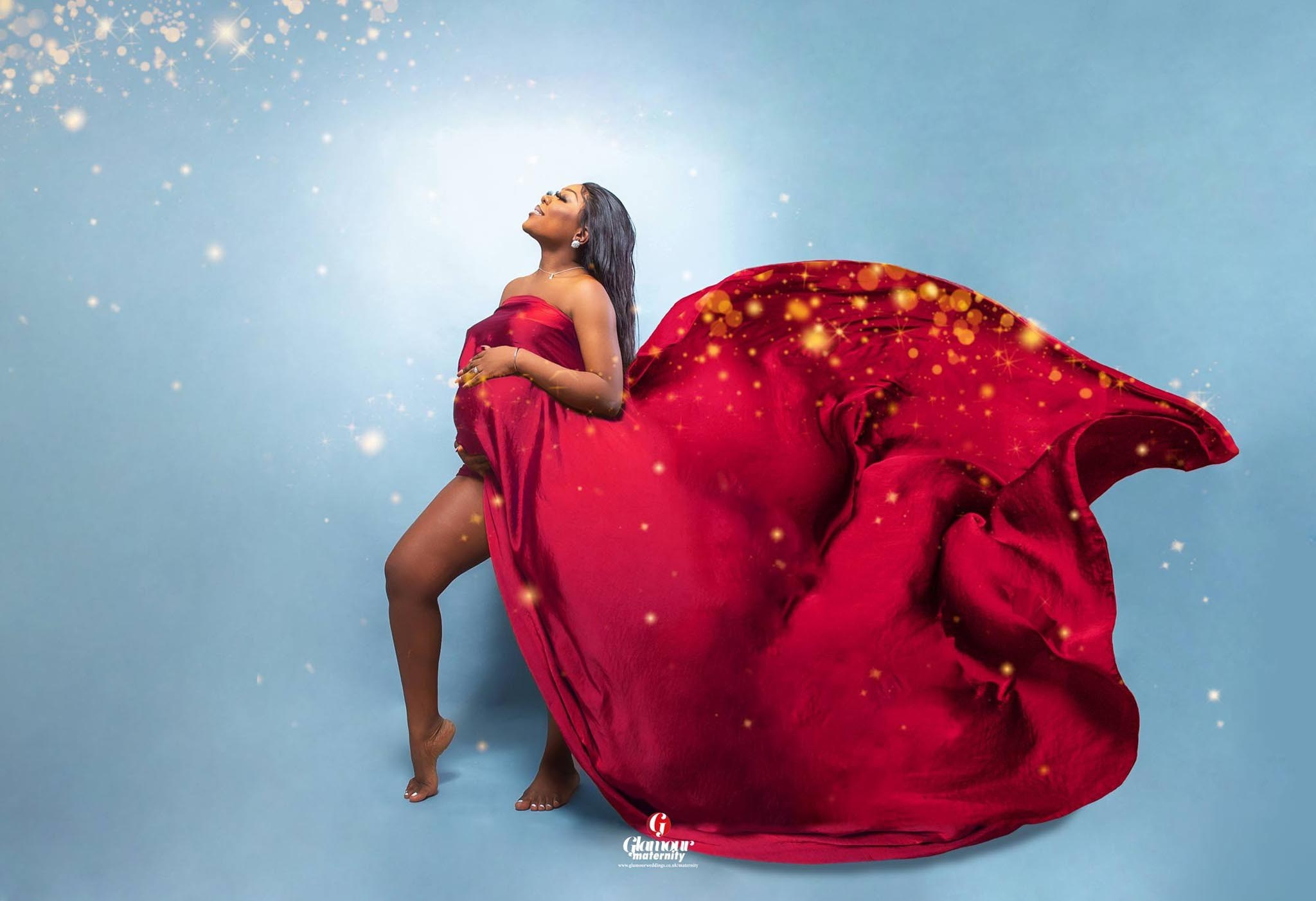 Creative maternity portrait wrapped in dramatic red fabric with glowing light effects against a soft blue background.