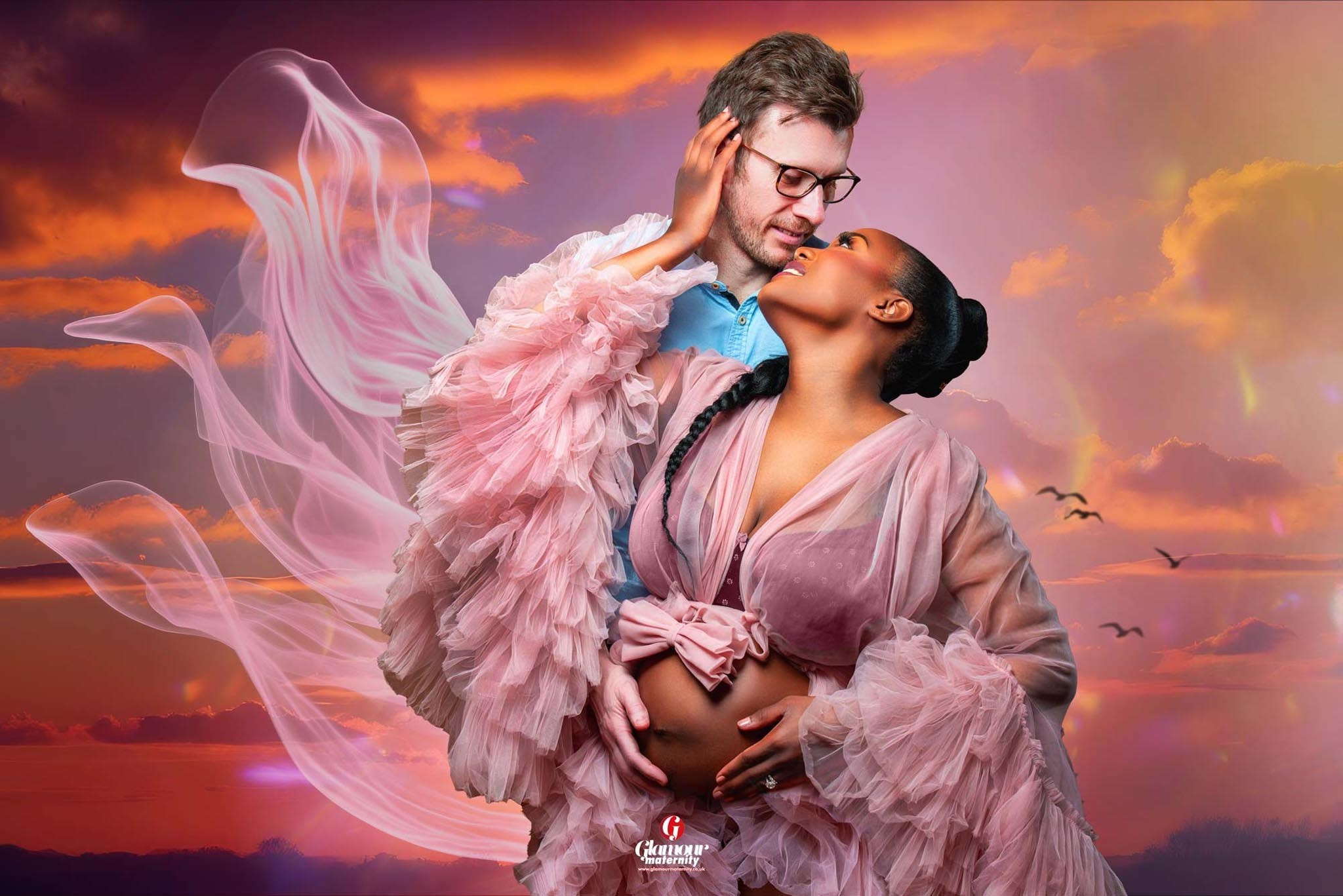 Intimate maternity portrait of an expectant couple in soft pink styling against a dramatic sunset-inspired background.