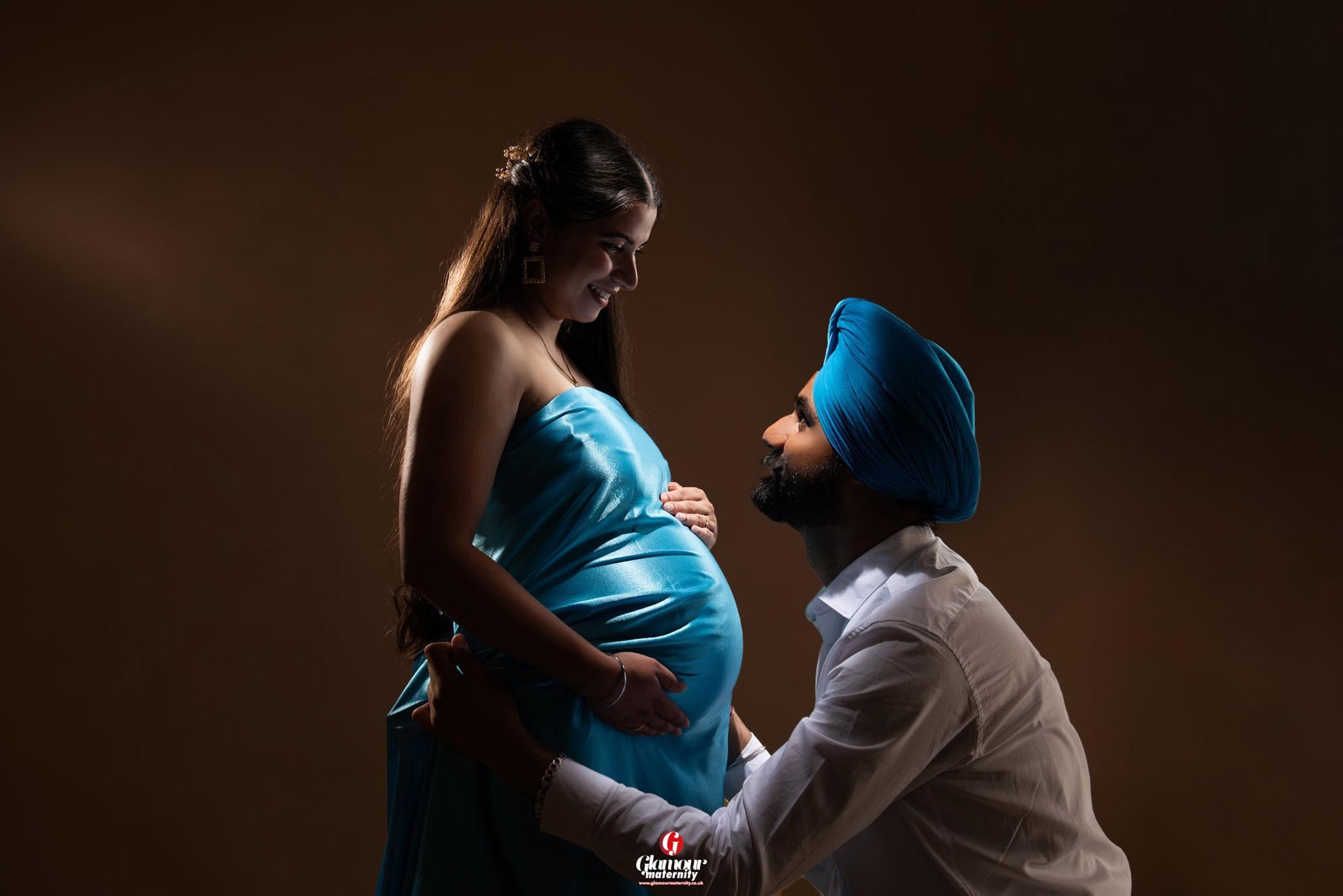 Tender studio maternity portrait of an expectant couple in blue styling, looking at each other in dramatic low-key lighting.