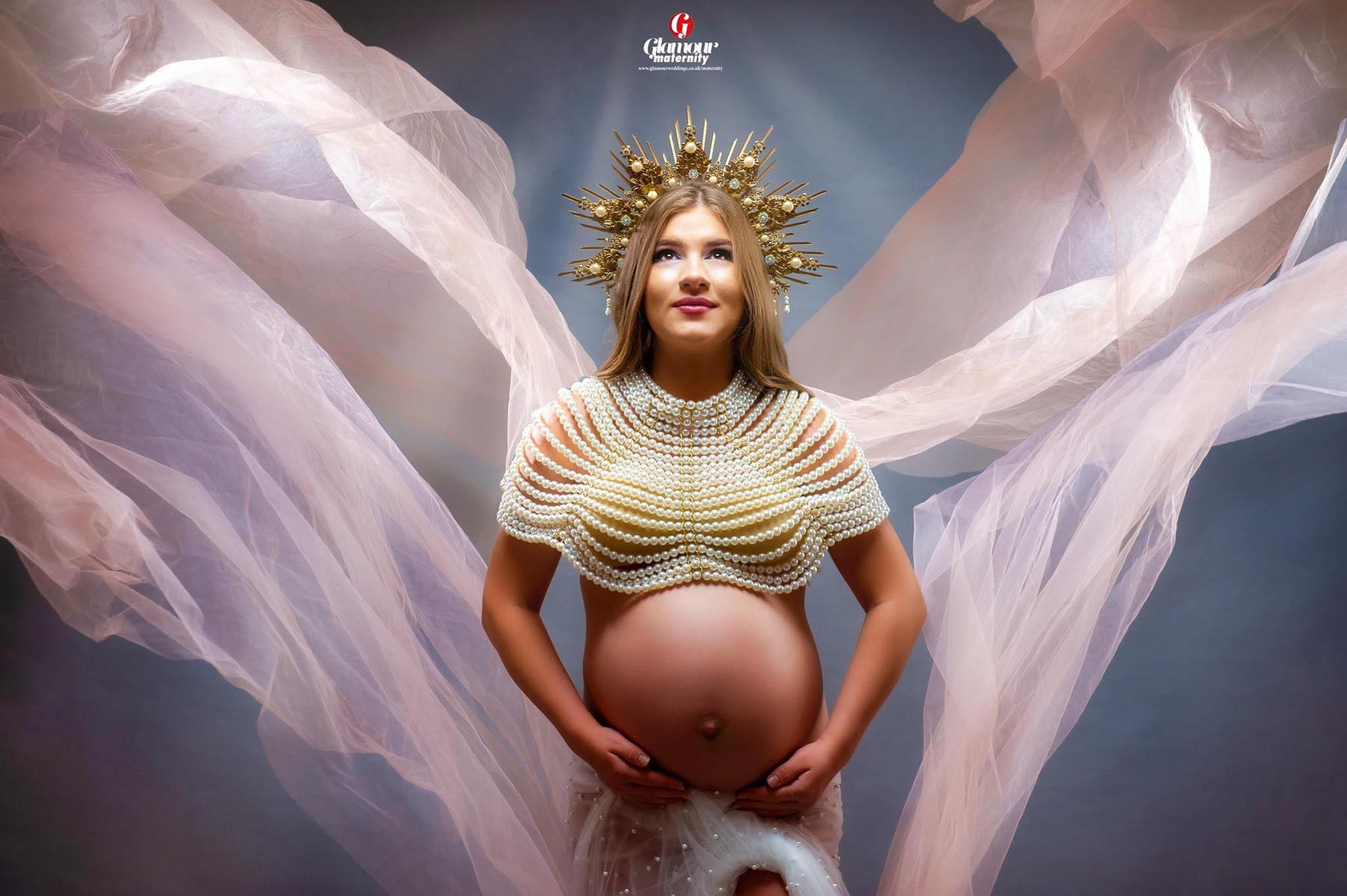 Fine-art maternity portrait with an ornate crown, pearl bodice and flowing sheer fabric against a soft grey background.