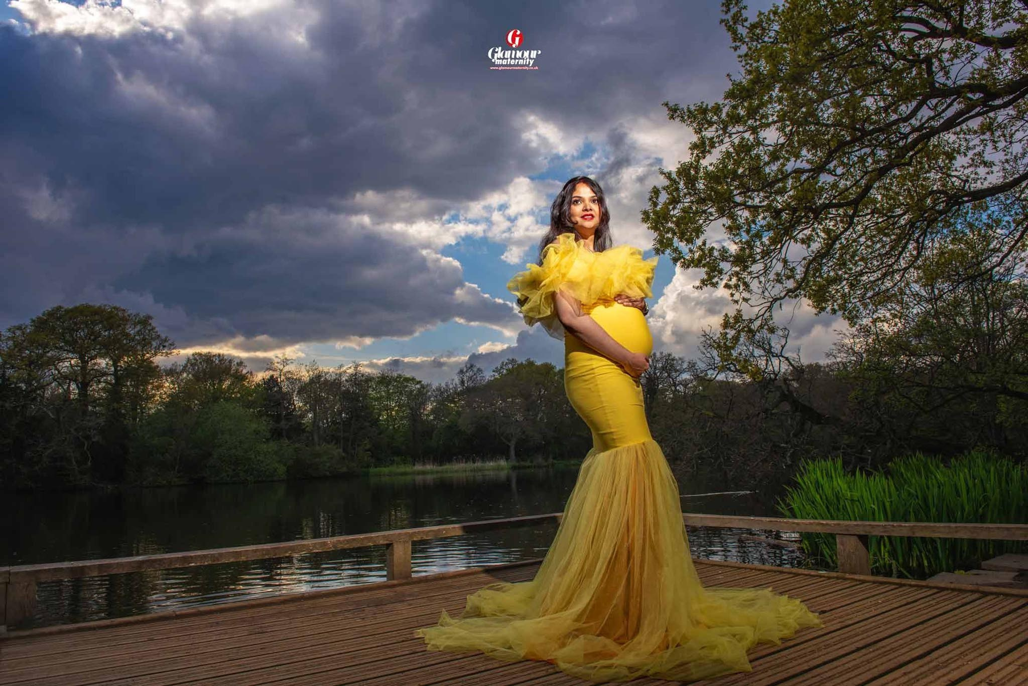 Outdoor maternity portrait in a flowing yellow gown beside a lake beneath a dramatic cloudy sky.