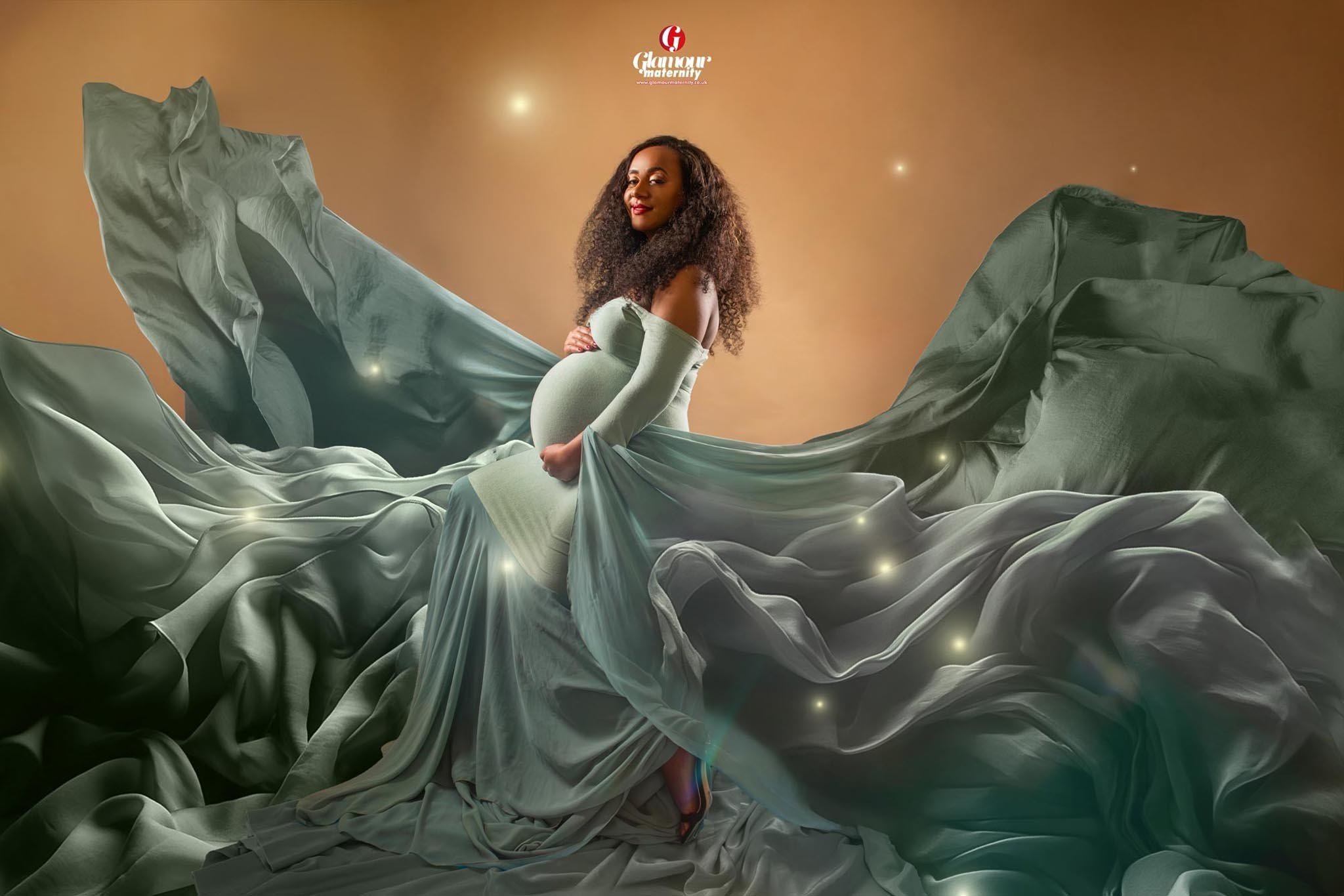 Glamour maternity portrait in a soft sage gown surrounded by dramatic flowing fabric and warm studio lighting.