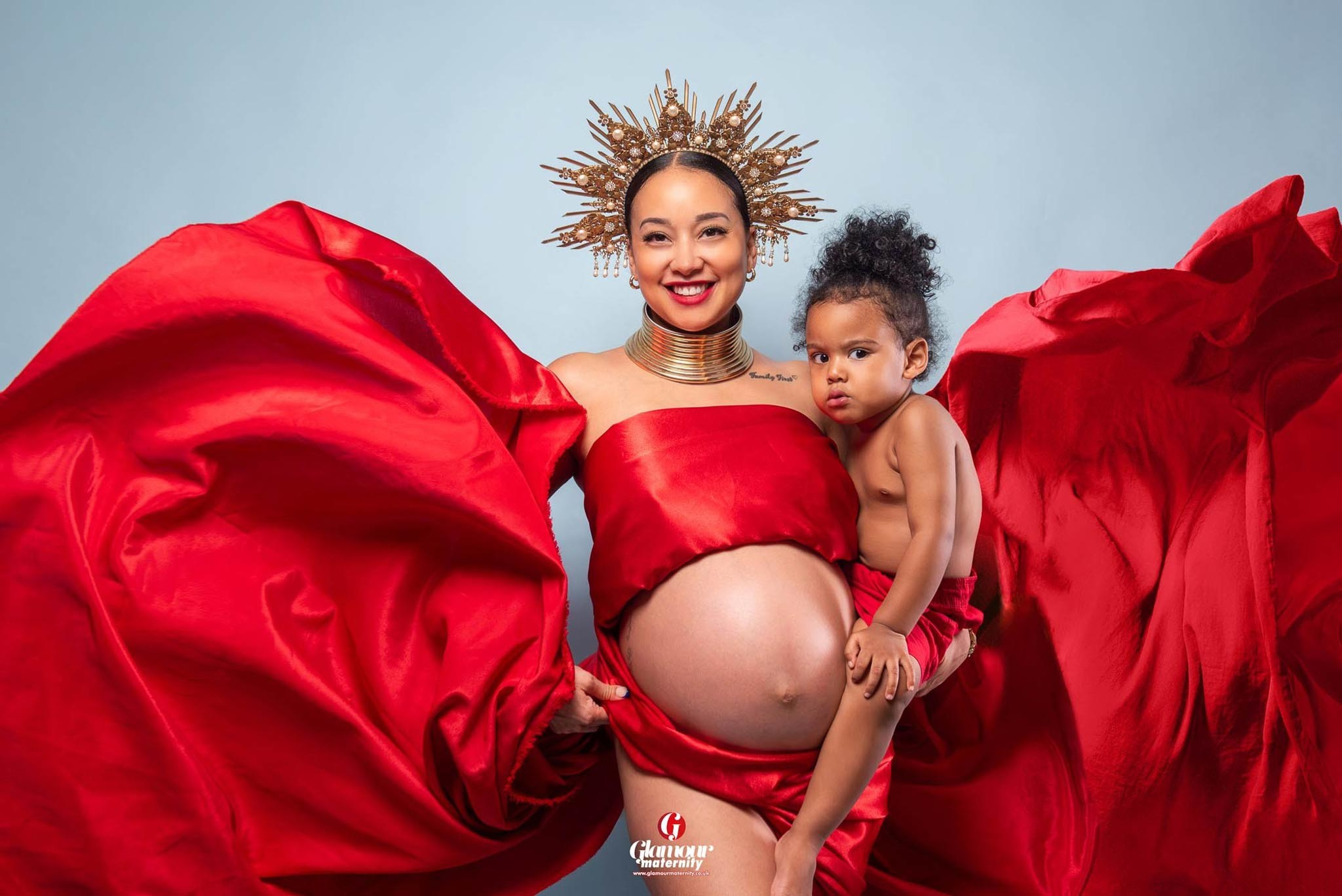Glamour maternity portrait of a pregnant mother holding her young child, styled with flowing red fabric and an ornate gold crown.