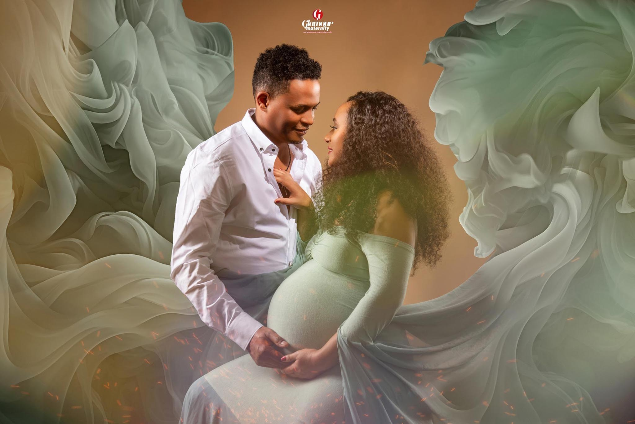Tender maternity portrait of an expectant couple in soft sage styling surrounded by flowing fabric and warm studio tones.