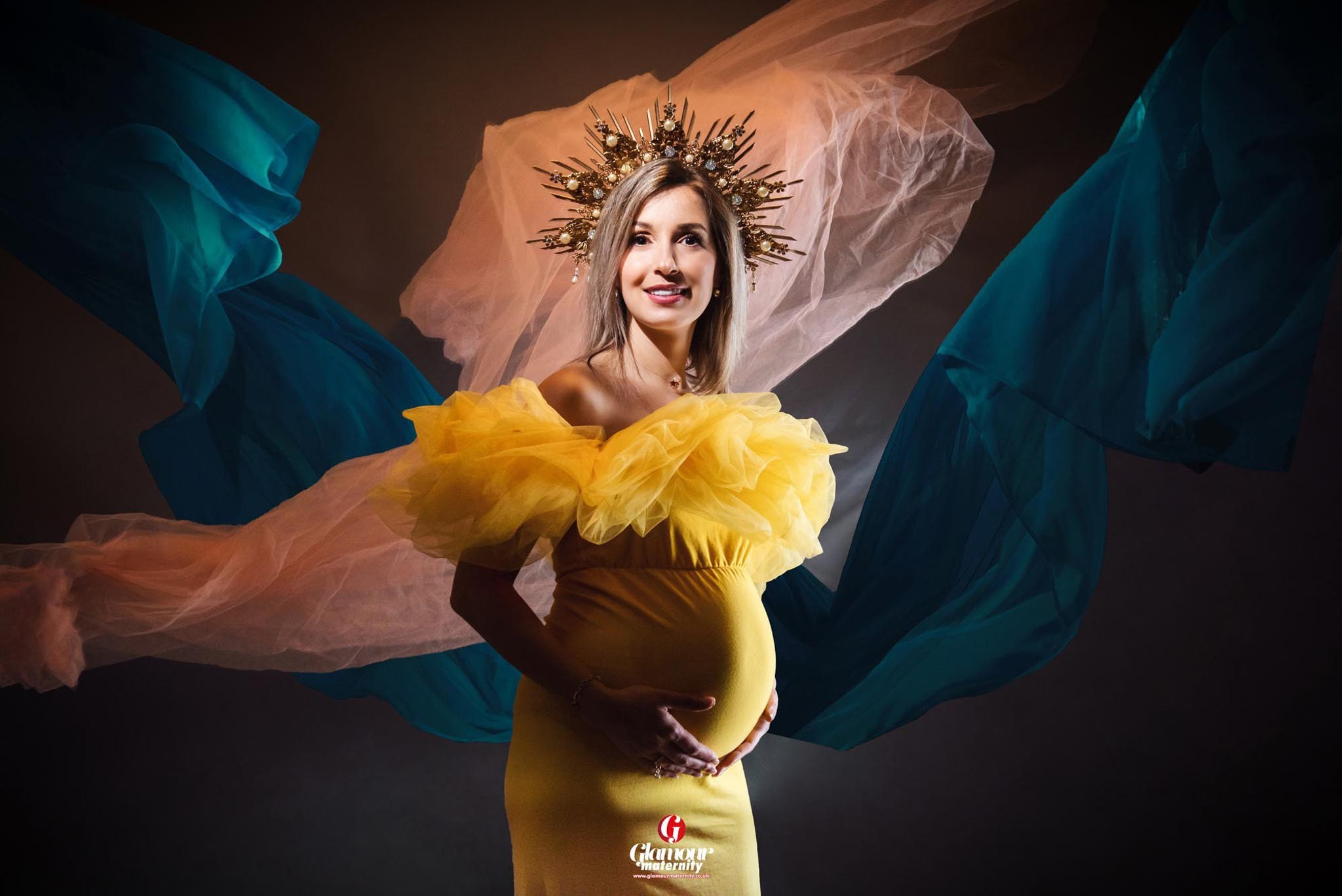 Glamour maternity portrait in a bright yellow gown with an ornate crown and dramatic flowing blue and pale fabric.